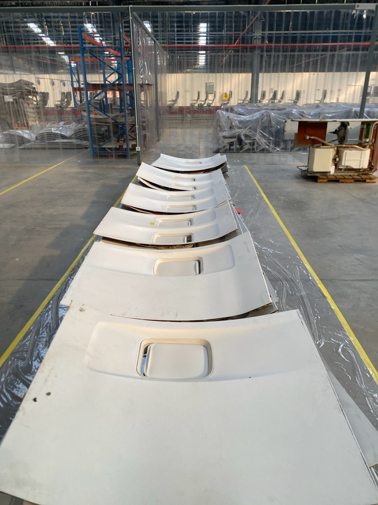 An overhead, long-shot, eye-level view captures a warehouse interior on Batam Island, Indonesia. In the foreground, a stack of white car hoods, each featuring a raised, recessed rectangular area and two circular indentations, are neatly arranged on plastic sheeting along a yellow painted line on the concrete floor. The hoods are stacked in a slightly offset, staggered manner, creating a diagonal line that recedes into the background. The warehouse is characterized by its industrial setting, with exposed metal beams and piping visible on the ceiling. Transparent plastic sheeting divides sections of the warehouse, revealing racks of industrial equipment and what appear to be chairs wrapped in plastic in the midground. In the background, further industrial equipment and storage units are visible. The overall scene suggests a storage or staging area for automotive parts within a manufacturing or distribution facility.
