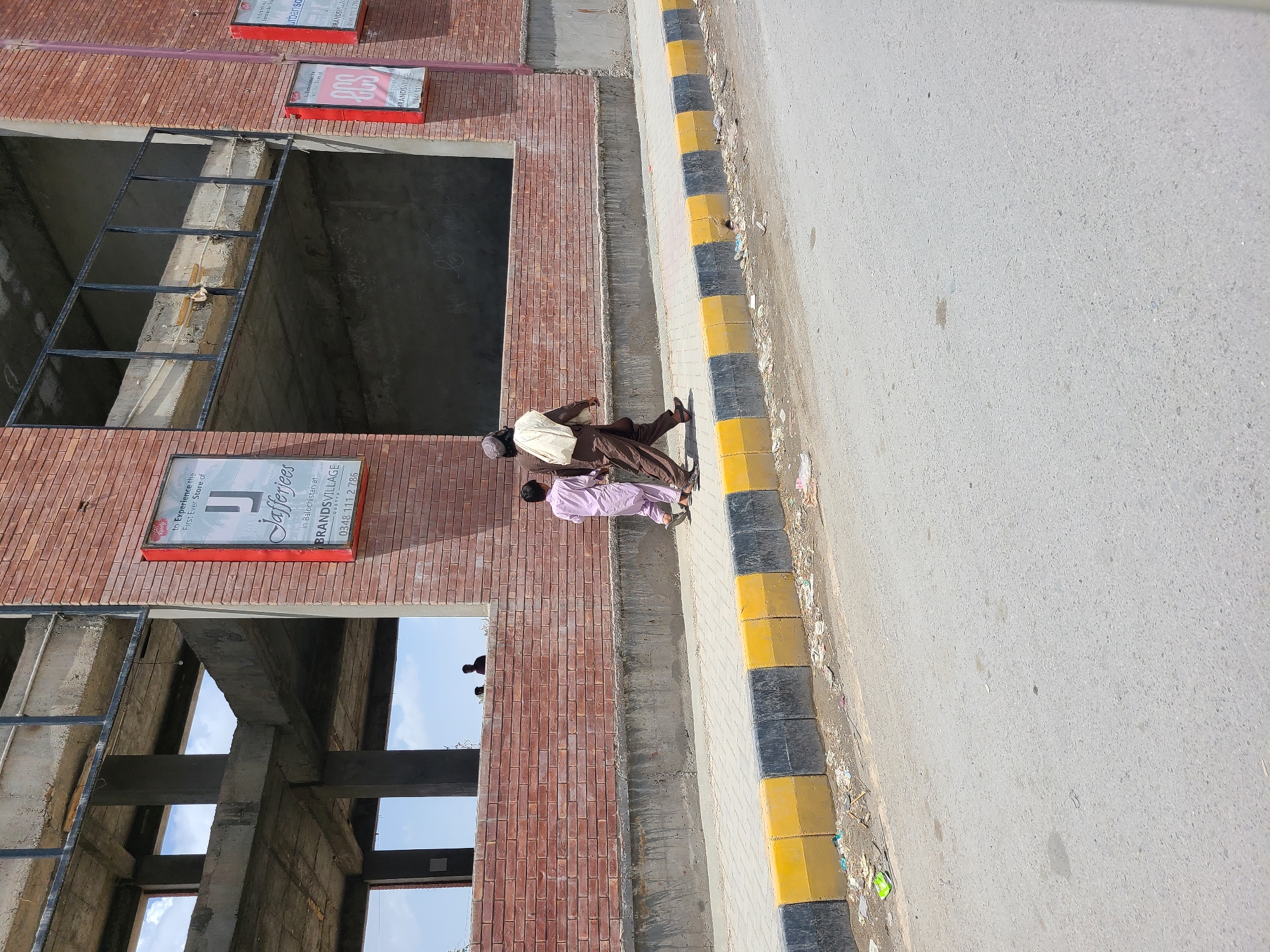 This media file captures an outdoor urban scene in Quetta City Tehsil, Pakistan, showing a building under construction alongside a street. The multi-story building features a facade of reddish-brown bricks with large, unfinished openings revealing concrete structural elements and metal framework within. Several red-framed advertising signs are attached to the brickwork. One prominent sign clearly reads "Jafferjees in Balochistan at BRANDSVILLAGE QUETTA" with a phone number "0348 111 2 786," and also includes text like "to Experience the First Ever Store of." Another sign further up is partially visible, showing "urbanso" and "2CS."
Two men are walking on the paved sidewalk adjacent to the building. One man is dressed in a light-colored shirt, dark trousers, and a dark cap, while the other wears a light purple traditional attire. The sidewalk borders an asphalt road, separated by a curb painted with distinctive alternating black and yellow blocks. Small debris and litter are visible along the roadside. The bright daylight indicates a clear, sunny day. Faint silhouettes of people can be seen inside the open structure of the building, possibly workers or onlookers. The image portrays a slice of everyday life against the backdrop of an ongoing commercial development.