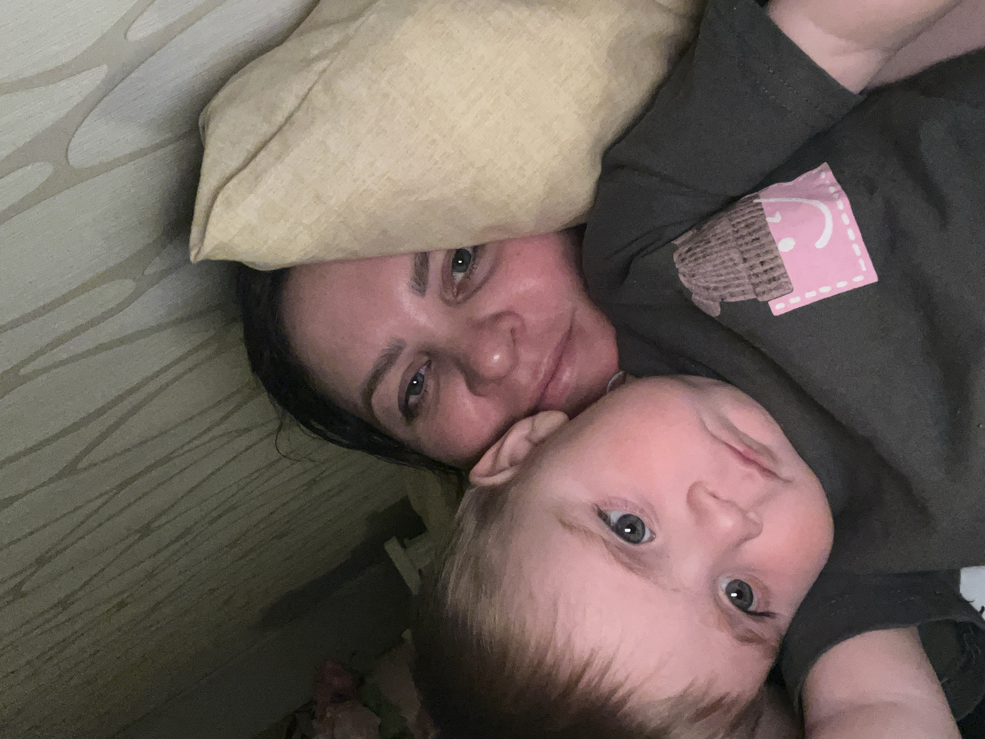 This media file captures a close-up selfie of an adult woman and a young child, likely a mother and her baby or toddler, lying together indoors. The image is taken from an overhead perspective, with both subjects looking towards the camera.
The woman, positioned with her head on a light-colored pillow, has dark hair and light eyes, displaying a gentle smile. The child, nestled close to her, has light hair and blue-gray eyes, looking directly at the camera with a curious, neutral expression. Both appear comfortable and content in the intimate setting.
The woman is wearing a dark-colored long-sleeved shirt. A notable detail on her sleeve is a distinctive patch or print: it features a brown, crinkled pattern resembling a cupcake wrapper, topped with a pink, rectangular section. This pink section has a white dotted outline and a white smiley face design. Behind the subjects, a wall with a subtle, light-colored wavy or abstract pattern is visible, consistent with an indoor environment. The lighting in the image is soft, suggesting a relaxed atmosphere. The provided context indicates the location is Perm, Russia.