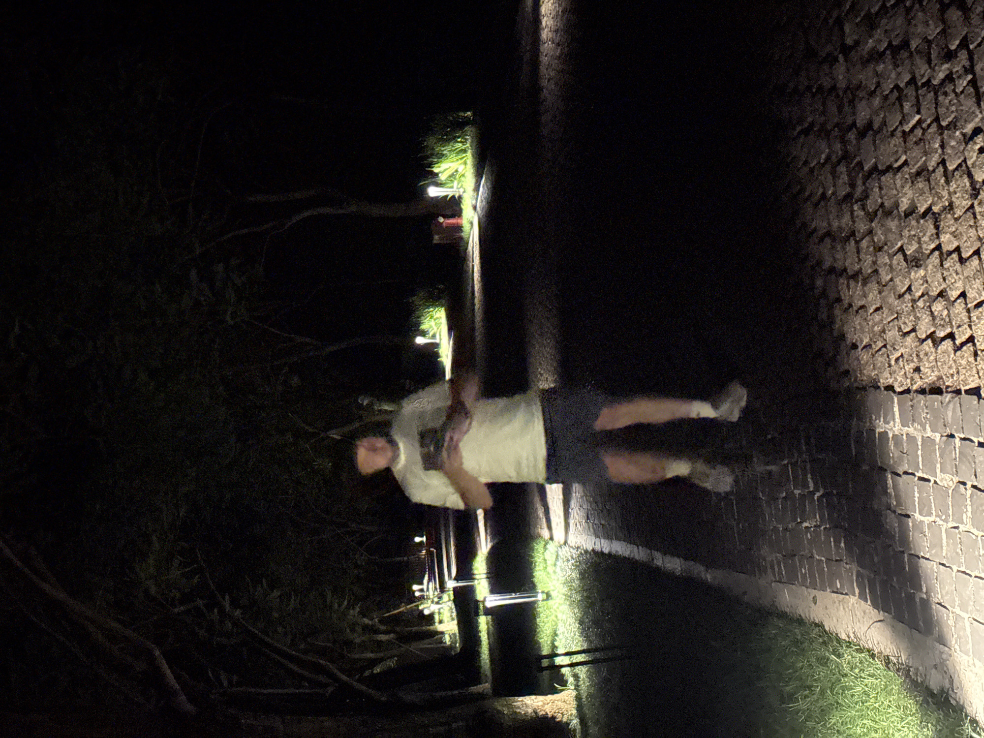 This image, taken at night in Mu Cang Chai, Vietnam, depicts a single male subject walking along a paved pathway. The subject, positioned centrally and moving towards the viewer, wears a light-colored t-shirt, dark shorts, light socks, and athletic shoes, and is holding a mobile device with both hands. The pathway, constructed from dark-colored paving stones, is lined on both sides by numerous ground-level lights that illuminate sections of the path and adjacent grass. Tall trees with dark foliage form a canopy overhead, creating a tunnel effect. In the middle distance on the right, a reddish cylindrical object is visible amidst brightly lit green foliage. The scene is characterized by deep shadows and concentrated pools of light from the artificial illumination.