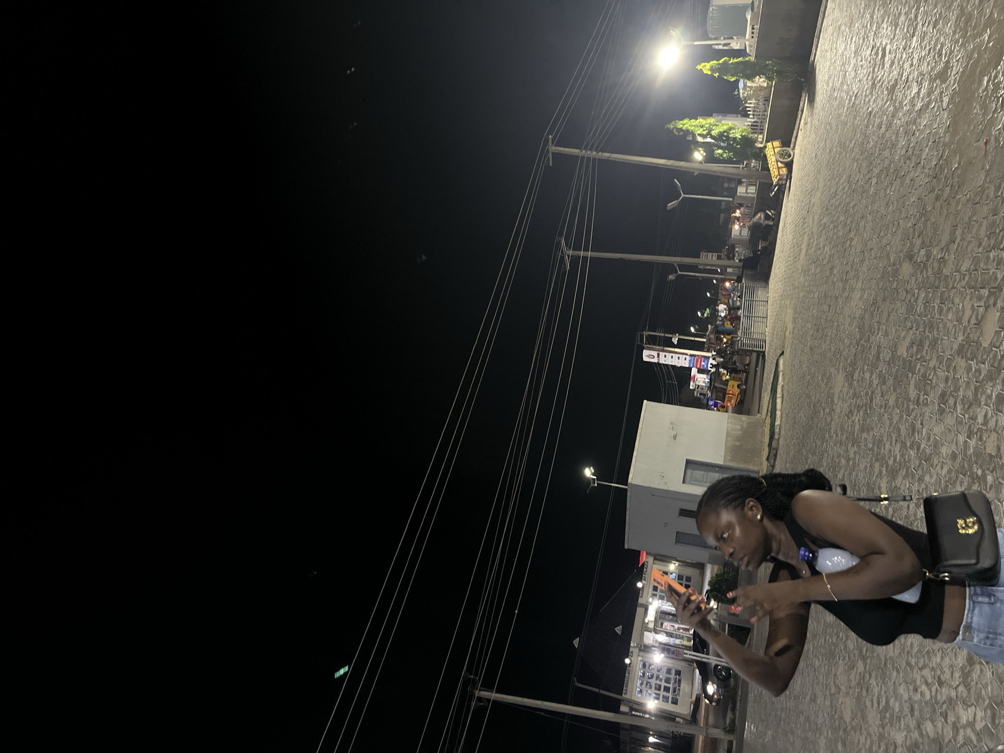 In Jalingo, Nigeria, at night, a Black woman with braided hair, wearing a black tank top and denim jeans, is taking a picture with her phone. She is holding a water bottle in her left hand and a black handbag with a gold buckle is slung over her right shoulder. The background shows a paved area with cobblestone patterns, streetlights, and buildings. Power lines are visible overhead, stretching across the dark sky. In the distance, there are signs for businesses, parked cars, and various vehicles, including a yellow cart with wheels. The overall scene depicts an urban environment at night, with artificial lighting illuminating the area.