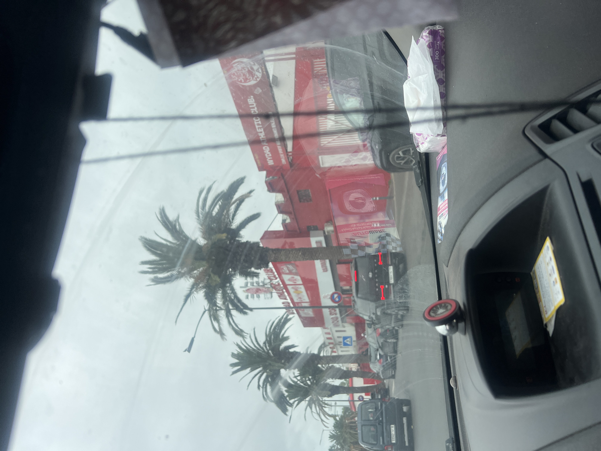 The image presents a view from inside a vehicle, looking out through the windshield onto an urban street in Casablanca, Morocco, on an overcast day. Reflections of the car's interior, including a tissue box and part of the dashboard with a circular dial, are visible on the glass.
Outside the vehicle, a prominent red building with white details stands along the street, featuring palm trees in front of it. Several cars are parked or moving on the road. A black SUV with illuminated red taillights is visible directly in front of the red building. Further down the street, another smaller dark vehicle is present. Road signs, including a blue circular sign with a red diagonal line and a blue square pedestrian crossing sign, are visible. The sky is uniformly gray, indicating cloudy or overcast weather.
Visible text includes: "WYDAD ATHLETIC CLUB:" prominently displayed on the red building, indicating a sports club. Other text fragments are "AWOTUA" and "VILLL BEE BOOM." On the tissue box reflected in the windshield, "300" and the Arabic word "رياضي" (Riyadi, meaning athletic or sports-related) can be seen. The overall scene captures a typical streetscape in a Moroccan city, with architectural and natural elements characteristic of the region.