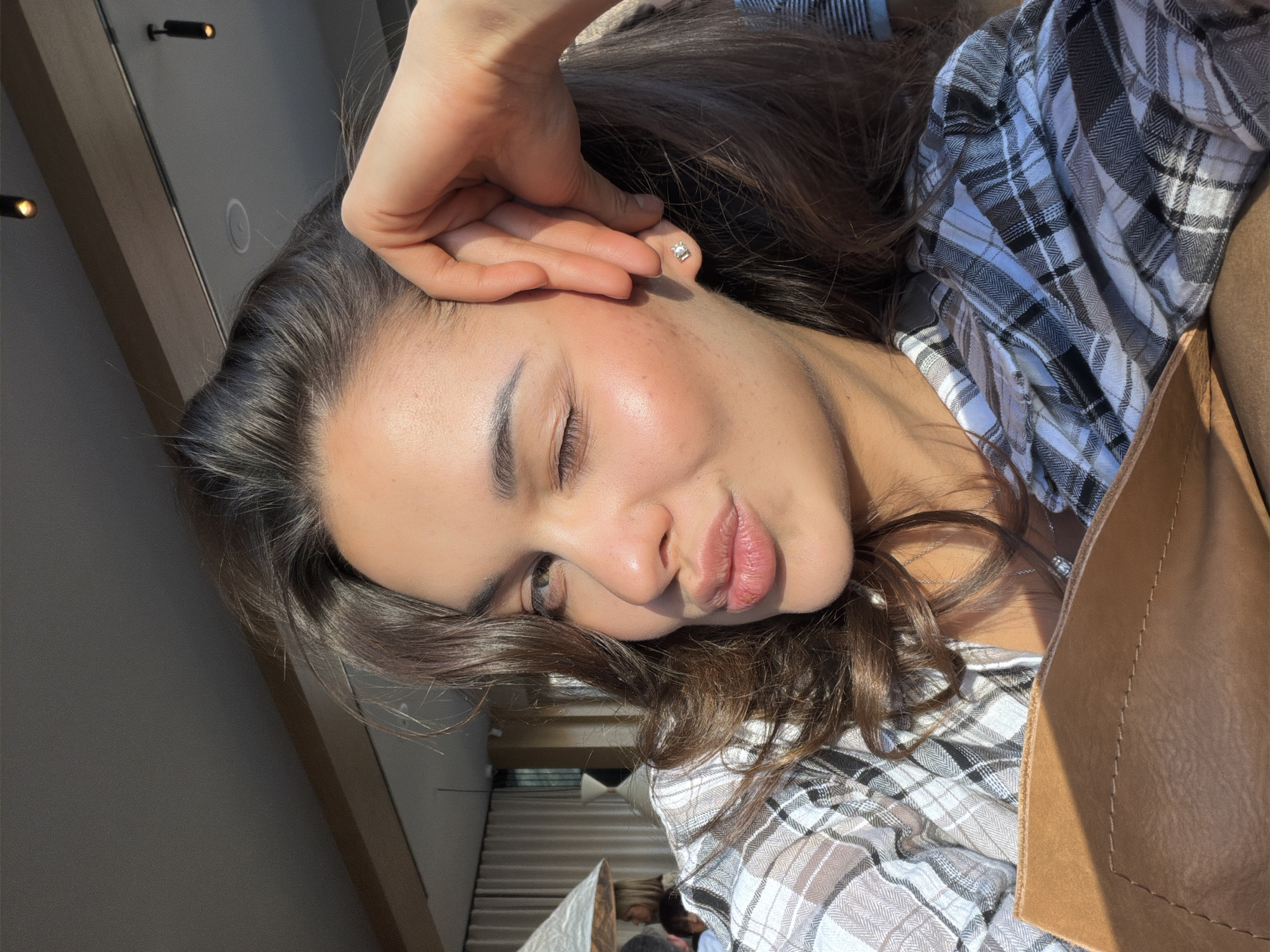 A medium close-up, selfie-style portrait features a young woman indoors. Her complexion is fair, with visible skin texture and a slight flush on her cheeks. Her dark hair is styled loosely around her face, and she has dark eyebrows and eyelashes. She wears a small stud earring in her left ear. Her eyes are closed, and her lips are slightly pursed. Her left hand rests on her forehead. She's wearing a gray and white checkered button-up shirt. A portion of a brown leather-like object appears in the lower right of the frame. The background is blurred and indistinct, with a partial view of a paneled wall and the possible suggestion of an interior space.
There is no verifiable information to definitively ascertain that this image was taken in Krasnoyarsk, Russia. The scene is generic and does not offer distinctive features to determine the location.