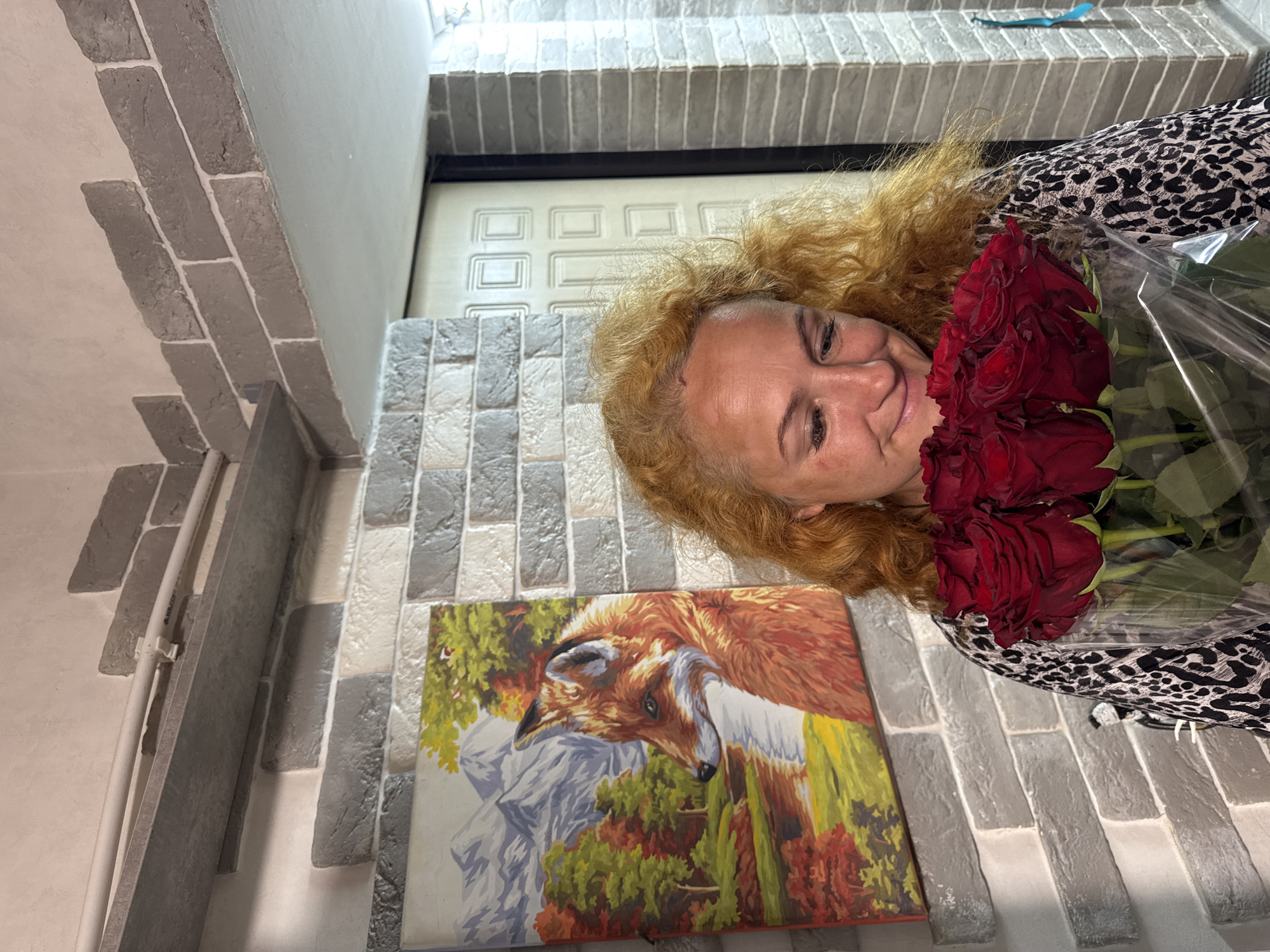 In the city of Chaikovsky, Russia, a woman with curly blonde hair and a leopard-print dress holds a bouquet of red roses. She is looking down at the roses with a smile. To her left, a painting of a fox in a natural setting hangs on a brick-patterned wall. The scene appears to be indoors, possibly in a hallway or entryway, with a door visible in the background. The lighting is natural and warm, casting soft shadows.