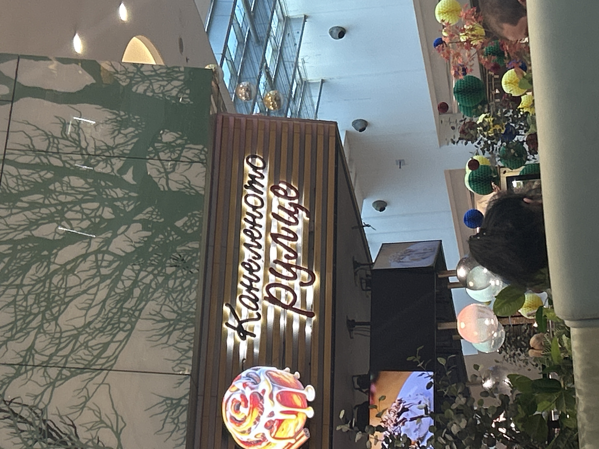 The image shows an indoor scene, likely a mall or shopping center. The focus is on a shop called "Канелено Руле" (Kaneleno Rule), which translates to "Cinnamon Roll" in Bulgarian. The shop's sign is vertically oriented, illuminated, and attached to a wooden panel. Below the sign is a colorful, stylized circular design, possibly representing a cinnamon roll. To the left of the shop, a wall is decorated with a print depicting tree branches.
In the background, the architecture reveals a multi-level building with a glass roof, allowing natural light to enter. Decorations, including colorful spheres and foliage, are suspended from the ceiling. People are present, although only partially visible, suggesting the scene is in a public, populated area. The scene is indoors. Due to the location being within Varna, Bulgaria, this is likely in a shopping mall or similar public indoor space in Varna.