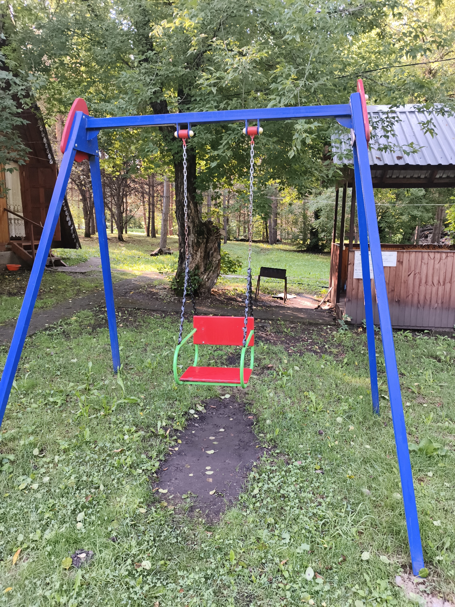 A front-facing, eye-level shot captures a child's swing set in an outdoor setting, likely in the city of Катунь, Russia. The swing set, constructed with blue metal poles, features a red seat with a green metal frame, suspended by metal chains. The ground beneath the swing is a patch of dark, damp earth with scattered fallen leaves. Surrounding the swing are areas of green grass with some weeds and patches of bare earth.
In the background, a wooden building with a dark roof and a small porch is visible on the left. To the right, a wooden structure with a corrugated metal roof and a wooden fence is present. Further in the background, a line of trees forms a dense forest. Sunlight filters through the leaves, creating dappled patterns on the ground. No people are present in the image. The overall scene depicts a somewhat unkempt, natural environment with a playground element.