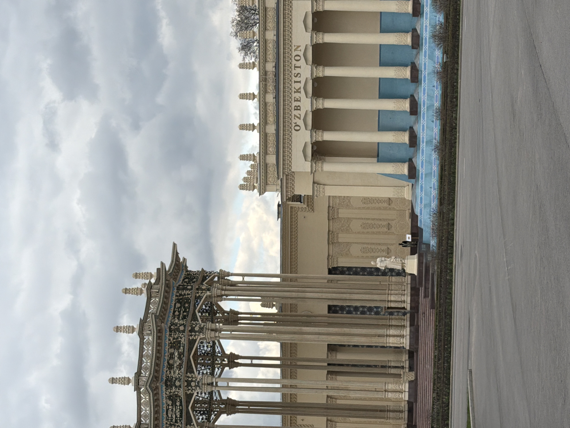 This media file captures a grand architectural complex, likely an exhibition pavilion, under an overcast sky. The scene is dominated by two ornate structures and an expansive paved area.
On the right, a prominent building features numerous light-colored columns and light blue sections along its facade, with intricate decorative patterns along the frieze and capitals. The word "O'ZBEKISTON" is clearly visible in large letters across the top of this building, identifying it as the Uzbekistan Pavilion. Bare tree branches are visible above its roofline.
To the left of this main building stands another highly decorative, open-sided structure, possibly a portico or a smaller pavilion. It boasts many slender columns and an elaborate, dark-accented roof with delicate patterns, suggesting a finely crafted design. Between these two structures, additional architectural details are visible, including more columns, decorative wall panels, and a white statue on a pedestal, possibly of a person, near what appears to be an entrance. A small flag or banner on a pole is also present.
The foreground and right side of the image show a wide, empty asphalt road or plaza. A narrow strip of green landscaping, featuring small bushes or trees, separates the paved area from the buildings. The sky is heavily clouded with light grey and white clouds, indicating an overcast daytime. No significant human activity is visible, contributing to a sense of quiet grandeur. Given the "O'ZBEKISTON" text and the location context, this is likely the Uzbekistan Pavilion at VDNH in Moscow, Russia.