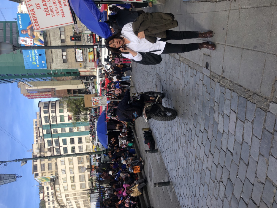 A woman stands on a brick sidewalk, talking on a cell phone. She wears a white blouse, dark pants, and brown boots, and carries a jacket and bag. Behind her, a crowded street scene unfolds with people lining the sidewalk and street. A motorcycle rider performs a wheelie amidst the crowd. Buildings rise in the background, interspersed with signs advertising "COMPRA Y" and the prices of dollars, euros, and pesos. Billboards promote Samsung and a mobile phone. The sky is partly cloudy.
