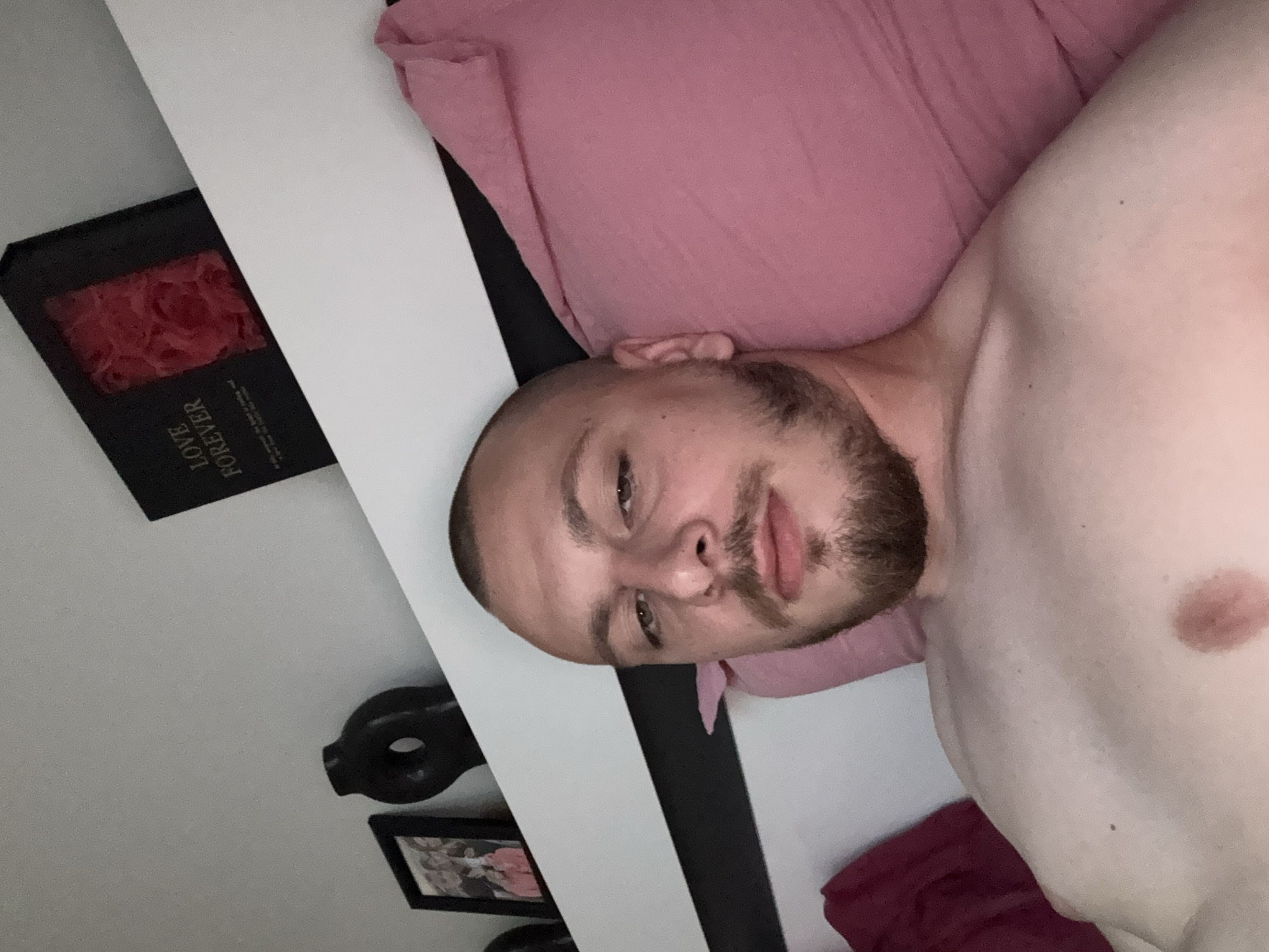 A man is lying down in bed, looking at the camera. He has a shaved head and a beard. He is wearing no shirt. Behind him, a pink pillow is visible. On a shelf to his left, there is a black box with red roses inside and the words "LOVE FOREVER" printed on it. Further down the shelf, there is a black decorative object and a framed picture. The scene appears to be indoors, in a bedroom. The lighting suggests it might be evening or nighttime. The man's expression is neutral.