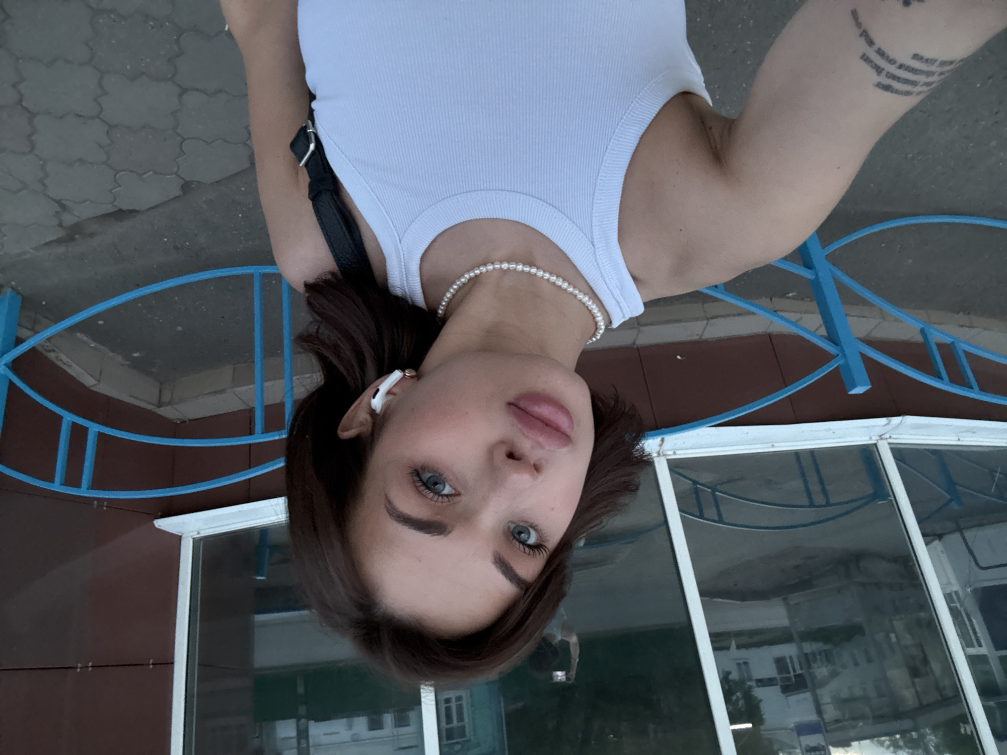 A selfie taken by a young woman with dark hair and light eyes. She is wearing a white tank top and a pearl necklace. She has a tattoo on her left arm and an earbud in her left ear. The background shows a bus stop structure with blue metal railings and a brown roof. The reflection in the bus stop windows reveals a building across the street. Based on the architectural style and available information, it is likely the image was taken in Sarapul, Russia.