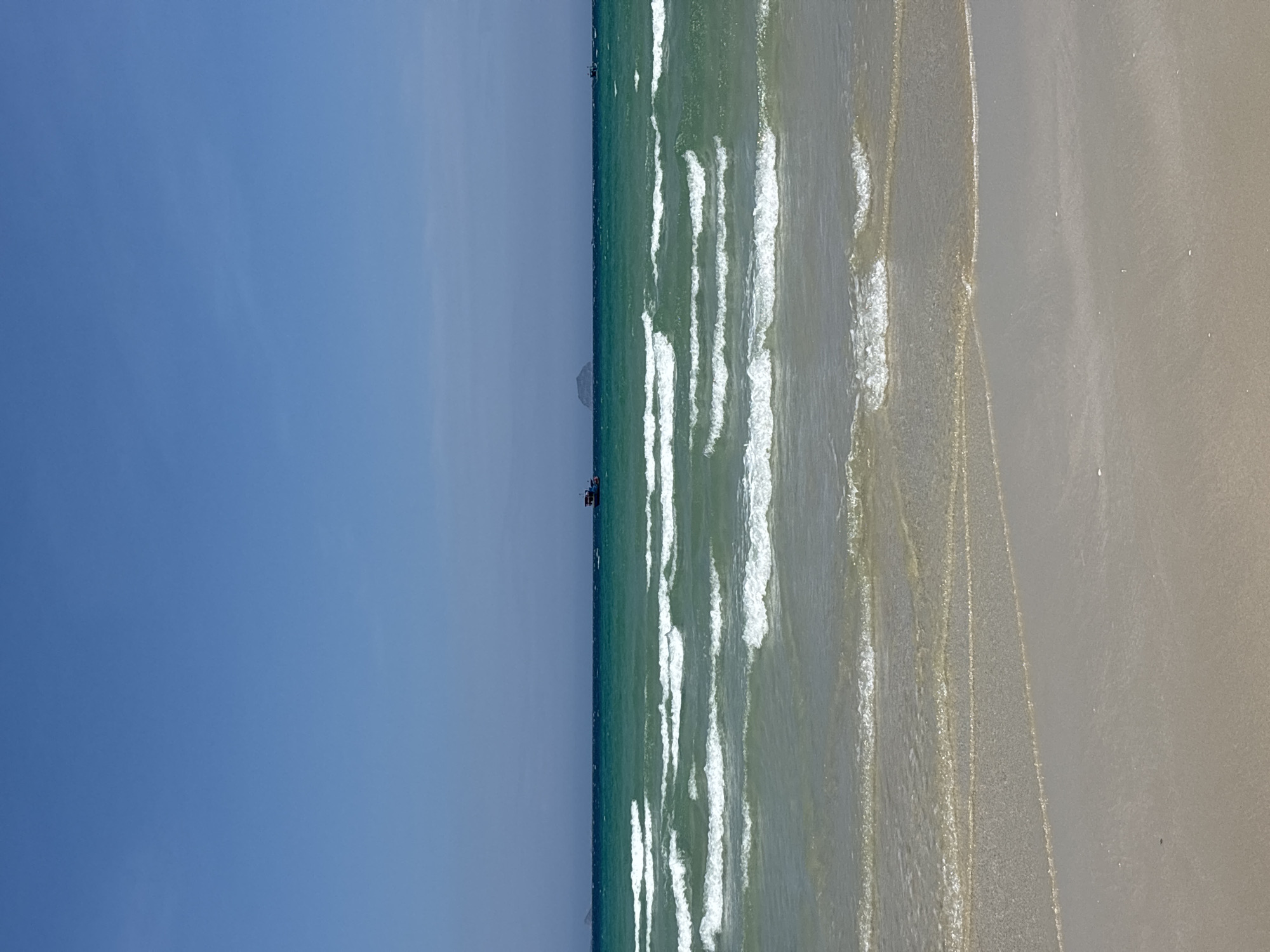 A wide shot of a beach shows a clear blue sky above a turquoise ocean. Gentle waves crash onto a sandy shore. In the distance, a small boat is visible on the water, and a distant island can be seen on the horizon. The sand is light brown and smooth, with some areas appearing wet from the receding tide. The water transitions from shallow and clear near the shore to a deeper, more vibrant turquoise further out.