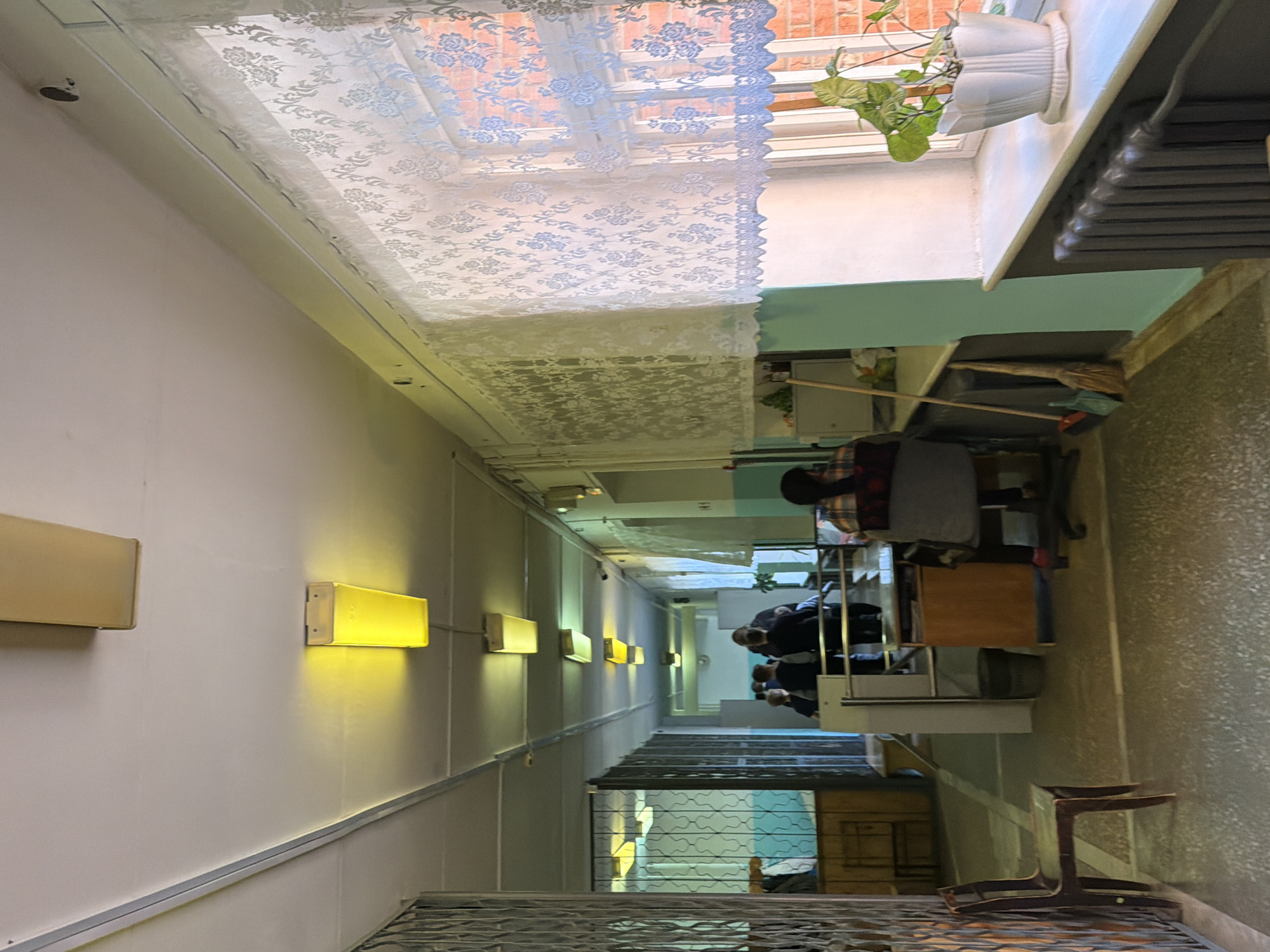 This image depicts a long, brightly lit indoor corridor or stairwell, likely within a public or institutional building in Noviy, Russia. The perspective looks down the length of the corridor, with a series of wall-mounted rectangular lights emitting a warm yellow glow along the left wall, illuminating the white surfaces.
Several people are visible in the middle distance and background of the corridor. They appear to be gathered, some seated at tables or desks, while others are standing. Their faces are indistinct due to distance and the lighting conditions. In the mid-ground, near where the people are, a broom and dustpan are leaning against a surface. A dark wooden chair with a distinctive, curved base is positioned in the right foreground.
At the upper part of the image (which would be the top of the corridor if viewed horizontally), there is a window covered by a white, semi-transparent lace curtain adorned with a floral or leaf pattern. Through the curtain, a red-brick building or wall is visible outside. A small potted plant with green leaves sits on the windowsill, and a dark radiator or heating unit is visible beneath the window. The strong artificial lighting suggests either evening or an interior space with limited natural light. There is no legible text visible in the image.
