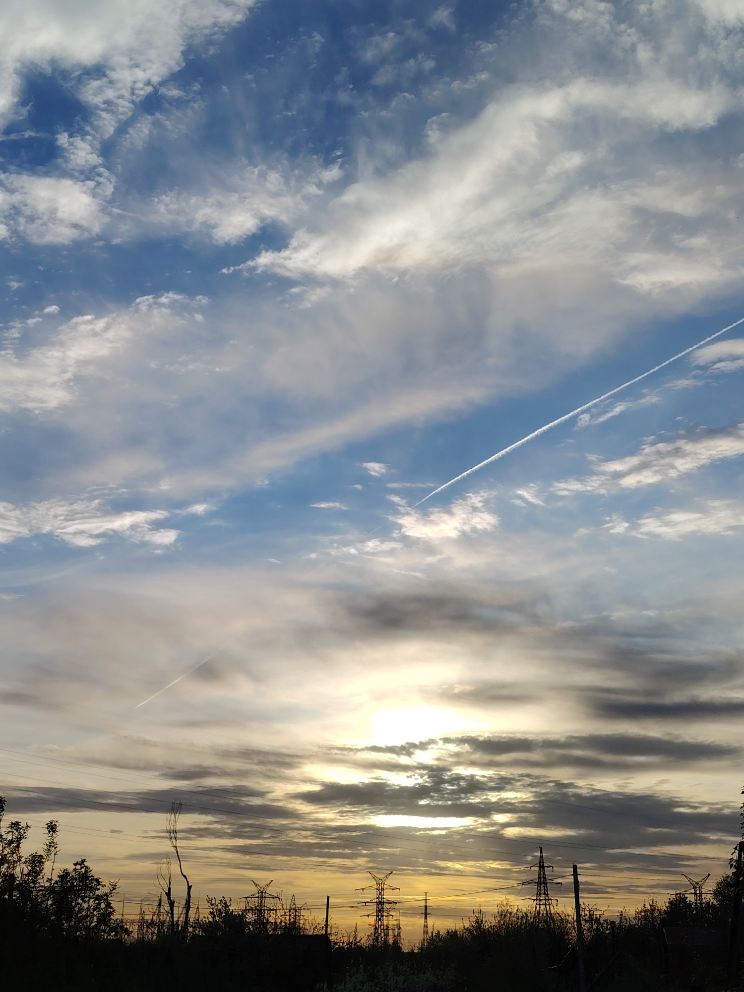 The image shows a sky with blue and white clouds, and two contrails. At the bottom of the image is a line of dark silhouetted trees and bushes, and some electrical pylons. The sun is obscured by clouds, but there is a bright glow. The location is in the city of Chaikovsky, Russia.