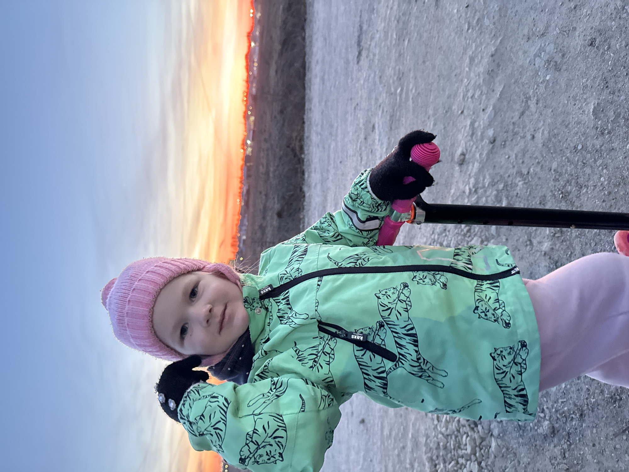 A portrait-oriented photograph, taken outdoors, shows a young child against a vivid sunset or sunrise backdrop. The image is originally rotated 90 degrees clockwise, depicting the child standing upright with the horizon to their right.
The central subject is a child, likely a toddler, looking directly at the camera with a calm expression. They are wearing a light pink knitted beanie, a light mint green winter jacket patterned with black cartoon tigers, black gloves with a silver or white embellishment on the back of the left hand, and light pink pants. The jacket features black zippers and a small tag with "KAXS ACTIVE" visible near the chest zipper.
The child is holding the pink and black handlebar of what appears to be a scooter or small bicycle with their gloved hands. The ground beneath them is a light grey, textured surface, possibly gravel or concrete. The background transitions from a clear, light blue sky on the left to a dramatic, bright orange and yellow horizon on the right, characteristic of either dawn or dusk. Along this vibrant horizon, there are small, distant lights or structures visible. The overall impression is an outdoor scene during the golden hour, likely in a park or open area.