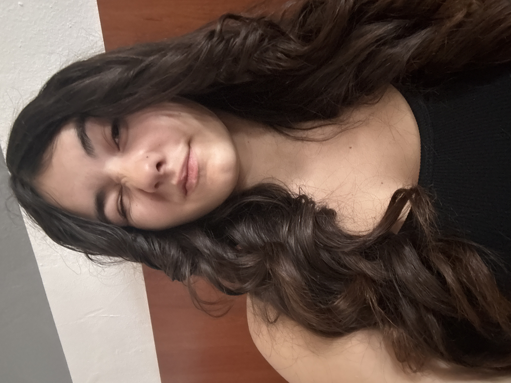 A woman with long, wavy dark brown hair is lying down, likely on a bed, wearing a black tank top. Her face is positioned towards the left of the frame, and she is squinting her left eye closed while smiling. The background consists of a wall with two distinct sections, one white/light gray and the other a reddish-brown. There are no indications that this image was taken in the city of Plovdiv, Bulgaria.