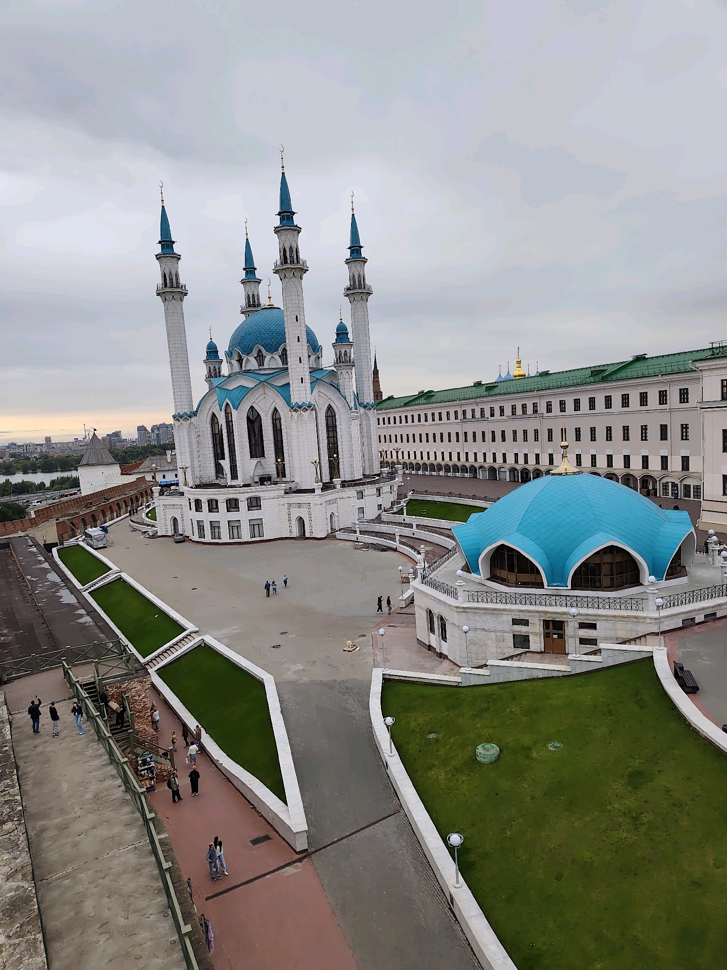 The image shows a high-angle view of the Kul Sharif Mosque in Kazan, Russia. The mosque features a prominent white structure with blue domes and minarets. Adjacent to the mosque is a smaller, also blue-domed building with arched windows. A long, multi-story building with a green roof and numerous windows runs behind the mosque. The courtyard area is paved with stone and features grassy areas and pathways. Several people are visible walking in the courtyard and on the elevated walkways. In the background, a cityscape with buildings and a body of water can be seen. The sky is overcast.