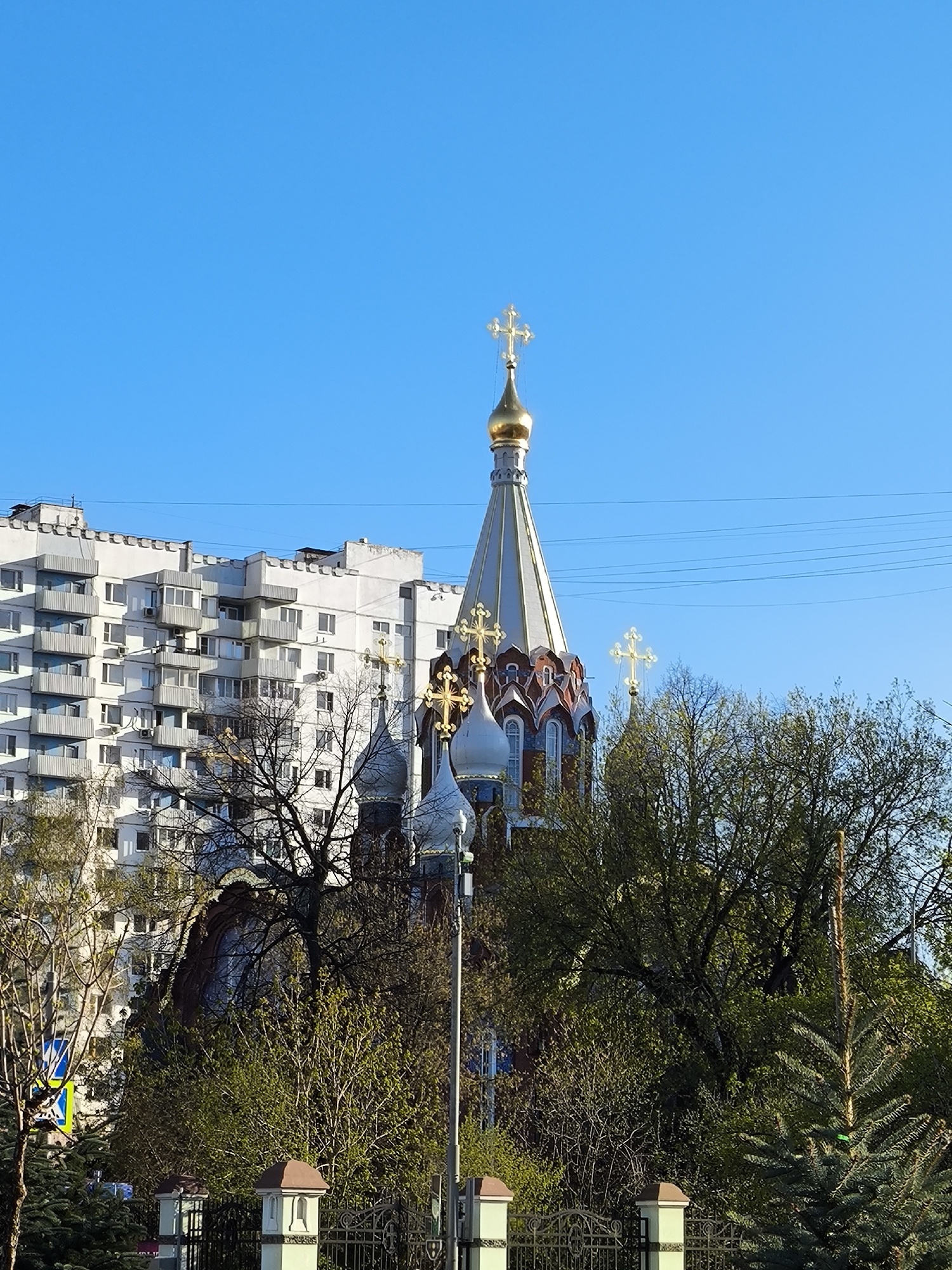 The image depicts a Russian Orthodox church in Moscow, Russia, partially obscured by trees and a fence. The church features a prominent golden dome topped with a cross, as well as smaller domes and crosses. Behind the church, a multi-story apartment building is visible. The sky is clear and blue. Power lines run across the upper portion of the frame. A section of a wrought iron fence with concrete posts is visible in the foreground.