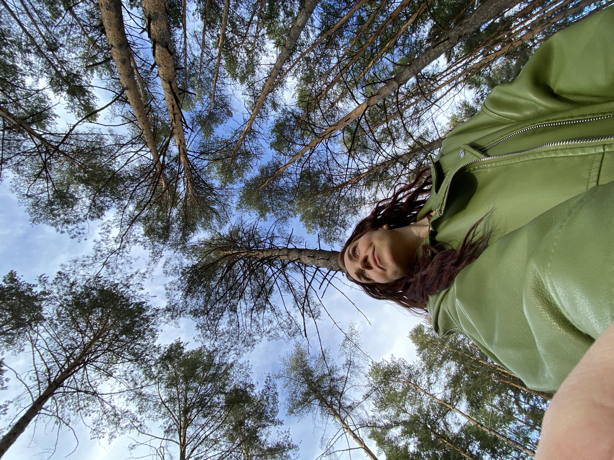 A low-angle shot looking up through tall trees towards a blue and cloudy sky. A woman is in the foreground, wearing a light green leather jacket. Part of her face and long dark hair is visible in the upper-right quadrant of the frame. The setting appears to be a forest, possibly within or near the city of Chaikovsky, Russia, given the prompt's requirement. The primary focus is on the trees and sky, creating a sense of depth and perspective.