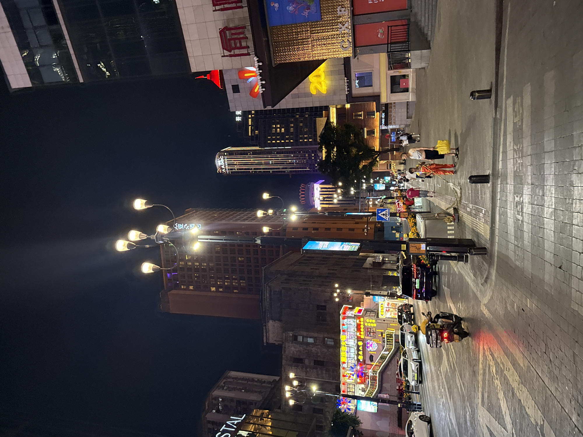 The image captures a nighttime street scene in the Yuzhong District of China. The street is paved with cobblestones and illuminated by streetlights and neon signs. Several buildings line the street, including tall, modern skyscrapers and older, more traditional structures. Cars and a scooter are visible on the road, suggesting moderate traffic. A few pedestrians are walking on the sidewalk, some carrying shopping bags. The overall atmosphere is one of urban vibrancy and commercial activity.
