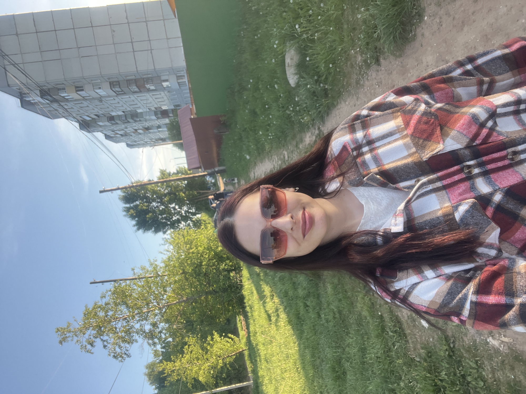 A woman wearing sunglasses and a plaid shirt stands outside in the city of Chaikovsky, Russia. In the background, there is a building, green grass, trees, and power lines. The woman is facing the camera.