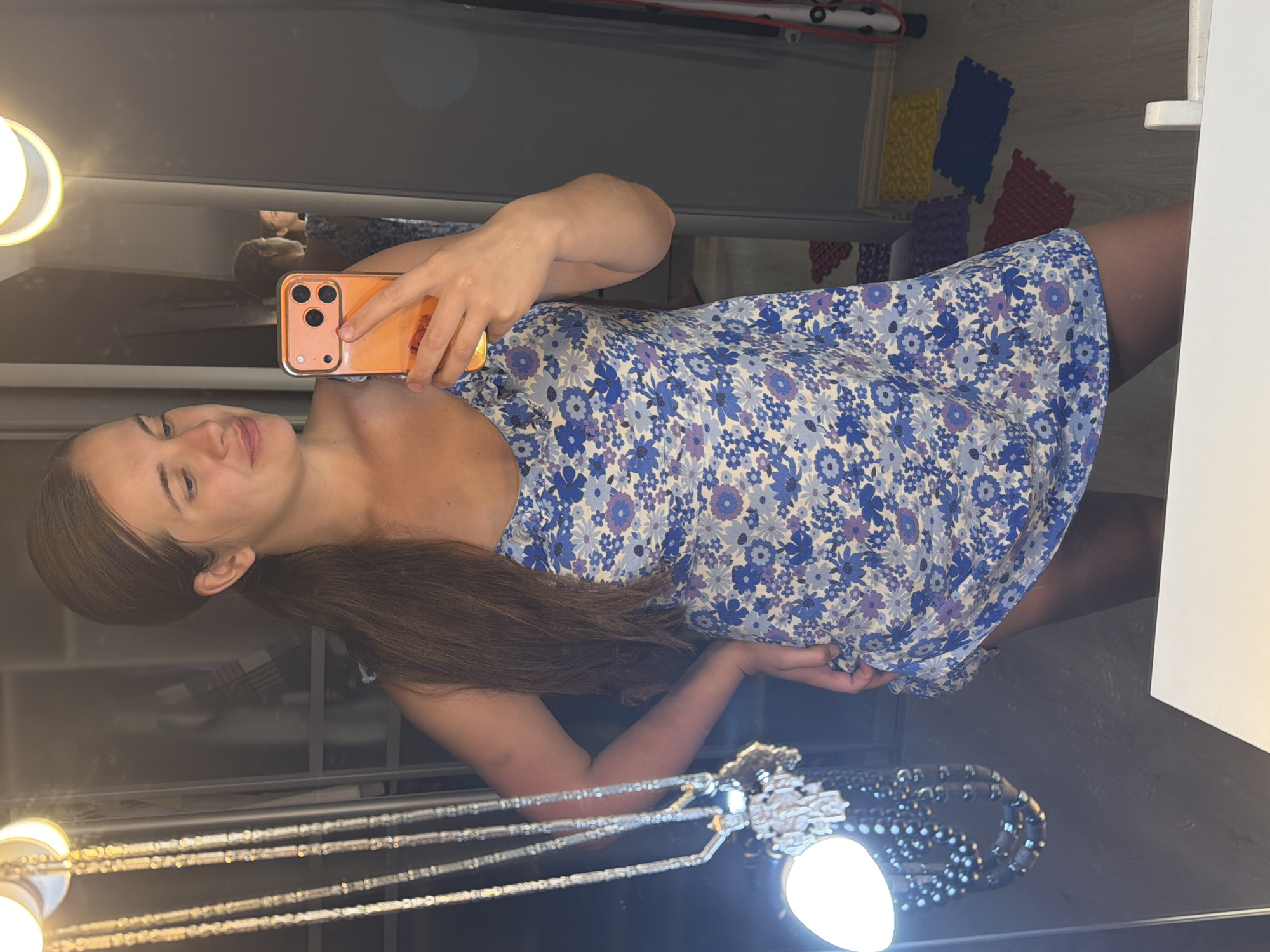 A young adult female, with long brown hair styled in a ponytail, is depicted in a mirror selfie taken indoors in the city of Sochi, Russia. She wears a blue, white, and purple floral print sleeveless mini-dress. She holds an orange-cased smartphone in her right hand, positioning it to capture her reflection, while her left hand rests on her hip. To the left of the frame, a tall stand holds multiple strands of silver-colored necklaces, some featuring intricate pendants, illuminated by a small spherical light source attached to the stand. Two additional circular wall-mounted lights emit a warm glow above this display. The background, visible through the mirror's reflection, includes a dark-colored wardrobe or closet with visible shelves and folded textiles. The floor is light-colored wood laminate, partially covered by several interlocking foam mats in blue, yellow, red, and purple. A red strap or band is also visible vertically near the mats on the right side of the reflected image.