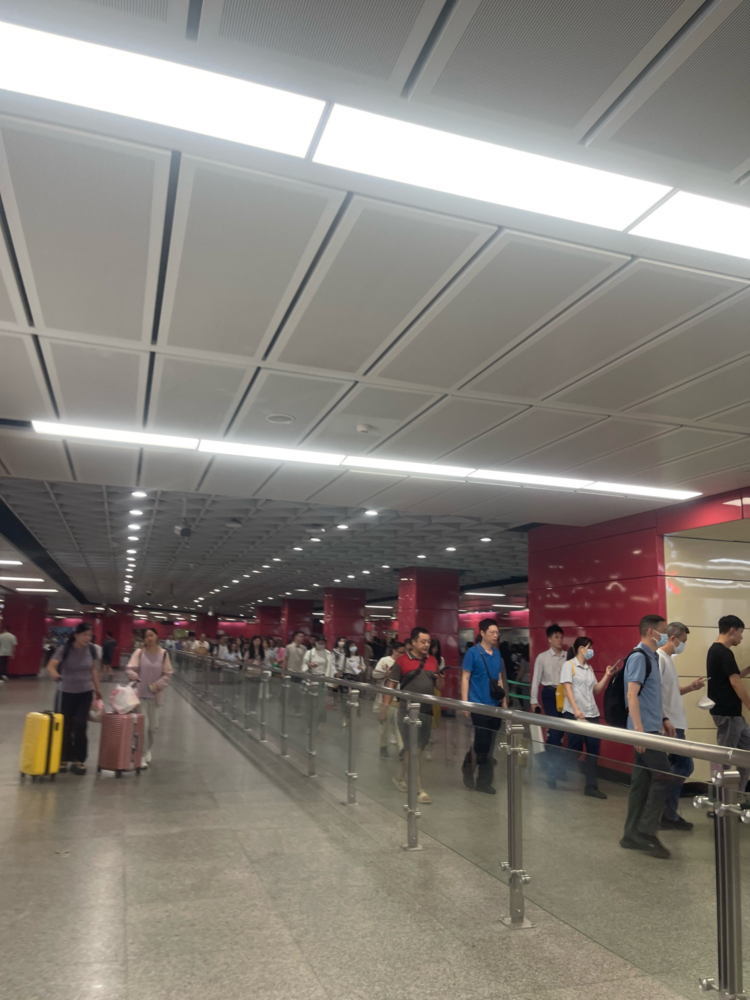 The image depicts a busy indoor public space, likely a subway station in Yuexiu District, China. The scene is characterized by a long, wide hallway with a polished concrete floor and bright, modern lighting. The ceiling is a suspended grid of acoustic panels with integrated fluorescent lights. On the right side of the hallway, a red wall paneling is visible, and a glass partition with metal railings separates the walkway from an unseen area. A crowd of people are walking in the same direction along the hallway, with some carrying luggage, suggesting travel. The individuals are diverse in age and gender, and many are wearing casual attire. Some are wearing face masks, indicating adherence to health protocols. The overall impression is one of orderly movement and activity within a functional urban transit hub.