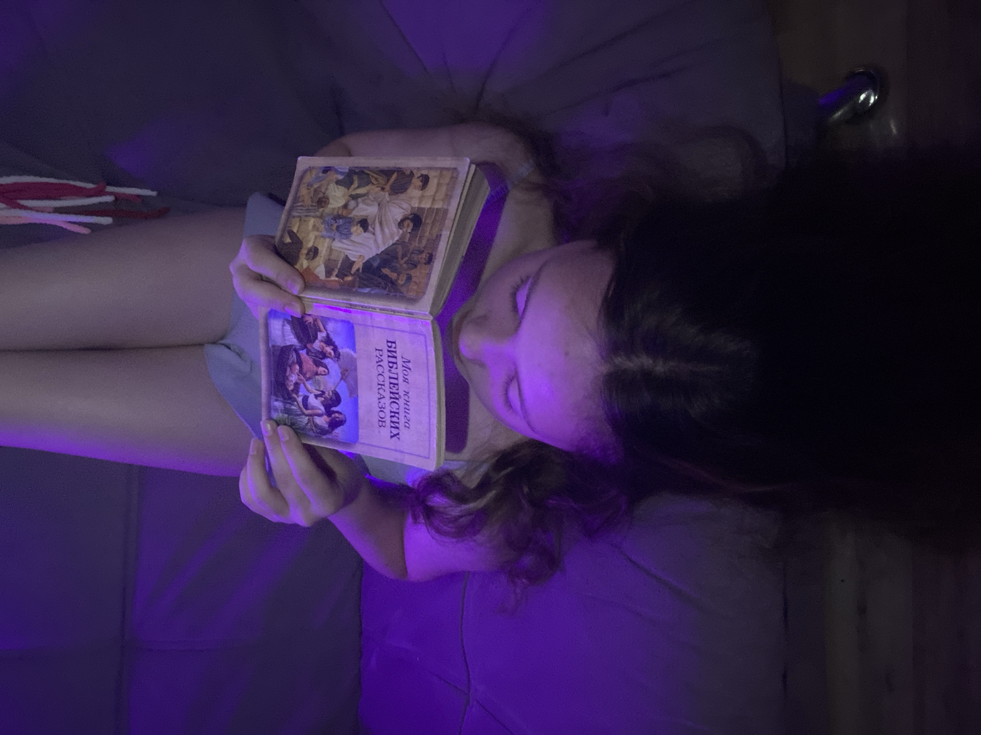 A person with long dark hair is lying down, possibly on a bed or couch, holding an open book in their hands. The book has a title in what appears to be Russian, and illustrations are visible on both open pages. The lighting in the scene is predominantly purple, creating a somewhat dark and moody atmosphere. The exact location is unknown, as is the gender of the person in the image. Other objects, possibly craft materials, are visible in the top left corner.