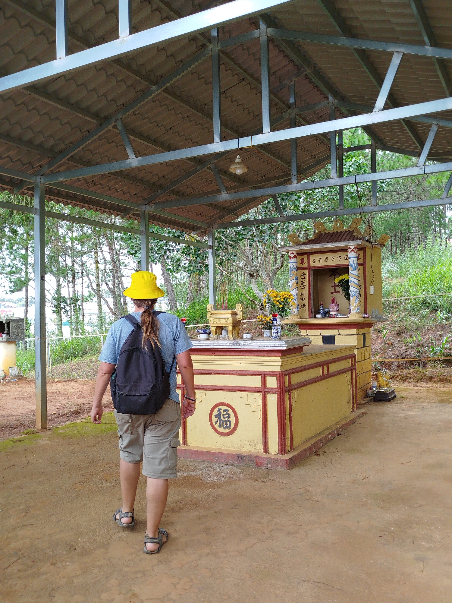 A person wearing a yellow bucket hat, a grey t-shirt, and khaki cargo shorts with a black backpack walks away from the camera towards a small, ornate, yellow shrine with a red roof. The shrine is decorated with blue and white Asian-style columns and has offerings placed on it, including yellow flowers. A large red circular symbol with Asian characters is painted on the side of a platform in front of the shrine. The scene is outdoors, under a metal-roofed structure with exposed beams, and surrounded by trees. The ground is a dirt or sandy path. The location is identified as Lam Vien - Da Lat, Vietnam.