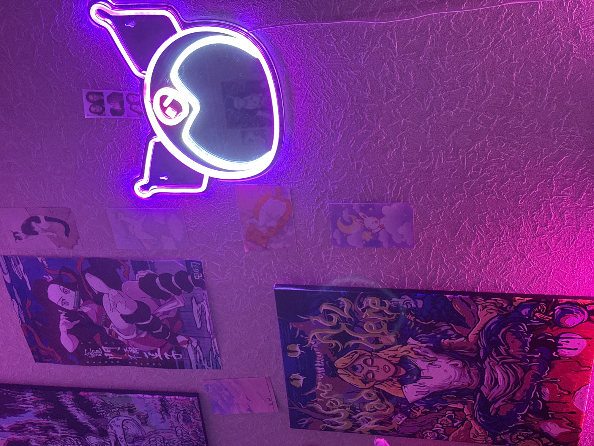 The image displays a wall adorned with various art pieces and a neon light fixture. On the left, a large vertical canvas depicts a figure in a meditative pose with swirling patterns and the words "We're All Mad Here". Above this, a smaller canvas features a surreal landscape with mushrooms. To the right of these, a poster of an anime character, Nezuko Kamado from Demon Slayer, is visible.
Further to the right, a neon light fixture in the shape of a pig's head glows brightly, casting a purple hue on the textured wall. Above the neon light, a strip of four small photo booth pictures are taped to the wall. Several other small art prints are scattered across the wall, including one with a crescent moon and a rabbit, and another abstract piece with orange and red hues. The overall lighting is a vibrant purple, suggesting a decorative or ambient lighting setup. The scene appears to be indoors, likely a bedroom or entertainment space.
