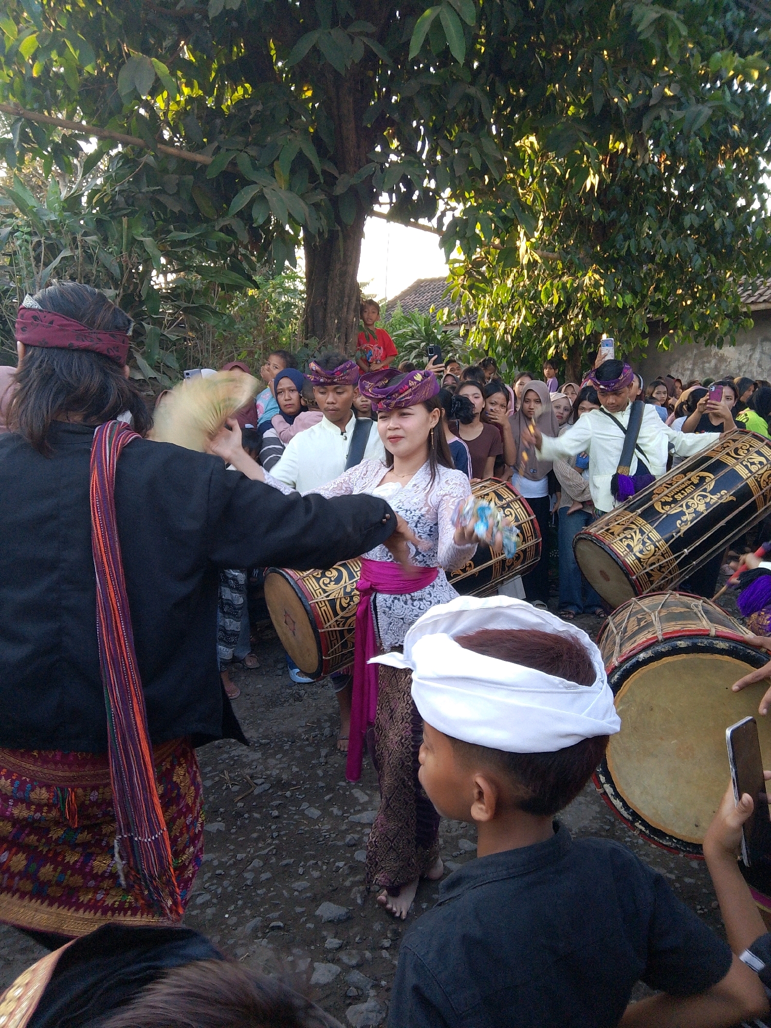 In Kuripan Utara, Indonesia, a group of people, likely participating in a cultural or religious event, are gathered outdoors. The scene features individuals in traditional attire, including women with decorative headbands and patterned sarongs. Several large drums, some elaborately decorated, are prominently displayed, suggesting a musical performance or procession. Some individuals are actively engaged in playing these drums, while others observe the proceedings. A young boy, wearing a white head covering, stands in the foreground, looking towards the activity. The environment appears to be a dirt or gravel area, with trees and a building in the background, indicating a village or community setting. The lighting suggests it is daytime, possibly late afternoon, given the warm tones.