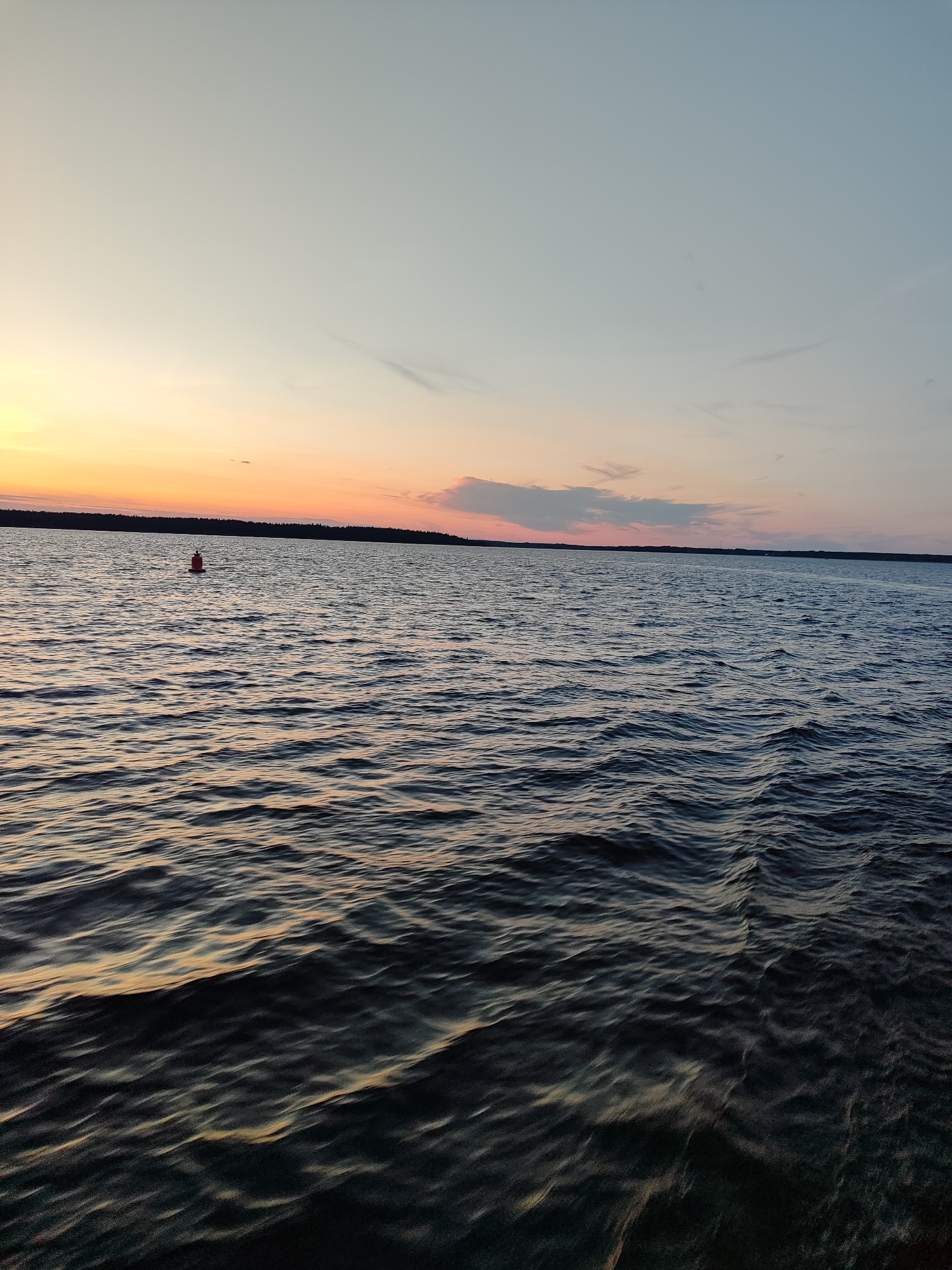 An outdoor, eye-level shot captures a calm body of water under a dusky sky. The water exhibits gentle ripples, reflecting the soft hues of the sunset. A single red buoy floats on the surface of the water, positioned slightly off-center to the left. In the distance, a dark, tree-lined horizon is visible, dividing the water from the sky. The sky transitions from a warm orange glow near the horizon to a pale blue and then a lighter blue towards the top of the frame. A few wispy clouds are scattered across the sky. The overall scene evokes a serene and tranquil atmosphere.
