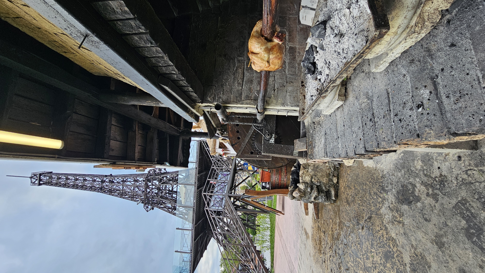 The image depicts a rotisserie chicken being cooked over an open fire, possibly in a restaurant or outdoor cooking setting. To the left, a miniature replica of the Eiffel Tower is visible. The tower is located in Varna, Bulgaria. The chicken is skewered and positioned above the fire. The setting is rustic with a mix of stone and wood construction visible.