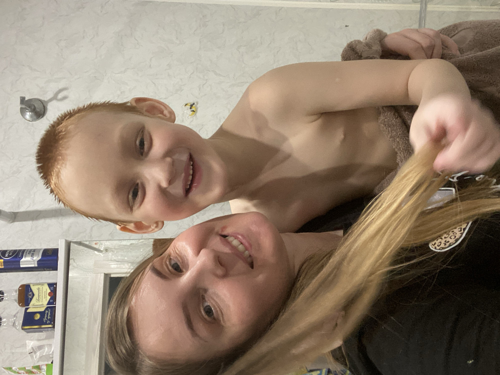 A woman and a young boy are photographed in a bathroom setting. The boy, with short red hair, is bare-chested and wrapped in a brown towel. The woman has long blonde hair. They are both smiling at the camera. The background includes a white tile wall, a shower head, and a shelf with toiletries and cleaning products. The location is indicated by the presence of "Noviy" in the product labels on the shelf, suggesting it is located in the city of Noviy, Russia.