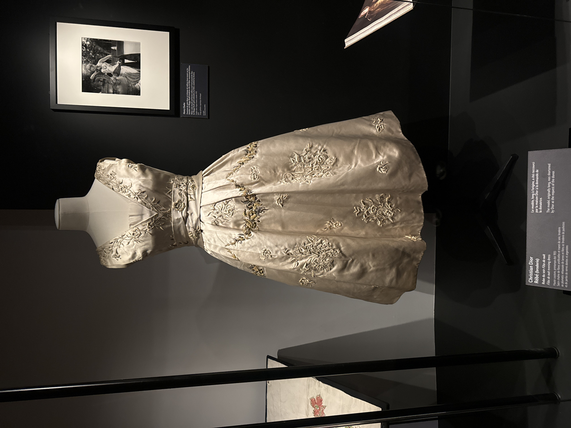 The image captures a display in what appears to be a museum or gallery, likely located in Paris, France, given the presence of French text. The focus is on a vintage, light-colored dress with intricate embroidery, displayed on a mannequin. To the left of the dress hangs a framed black and white photograph. A plaque next to the dress provides information in both French and English, indicating it is a "Christian Dior" evening dress titled "Fête de nuit" (Night Party), dating back to the Haute couture spring-summer collection of 1955. The plaque also notes the dress, originally long, was shortened by Dior at the request of the donor. Above and to the left of the dress is another plaque titled "Henry Clarke" providing further details, although the exact contents of that text are unreadable. The environment consists of dark walls and minimalist display stands, creating a muted backdrop to highlight the artifacts. The image appears to be professionally lit, with careful attention to the lighting of the dress. The scene has a modern and somewhat minimalist feel, befitting a contemporary museum or gallery space.