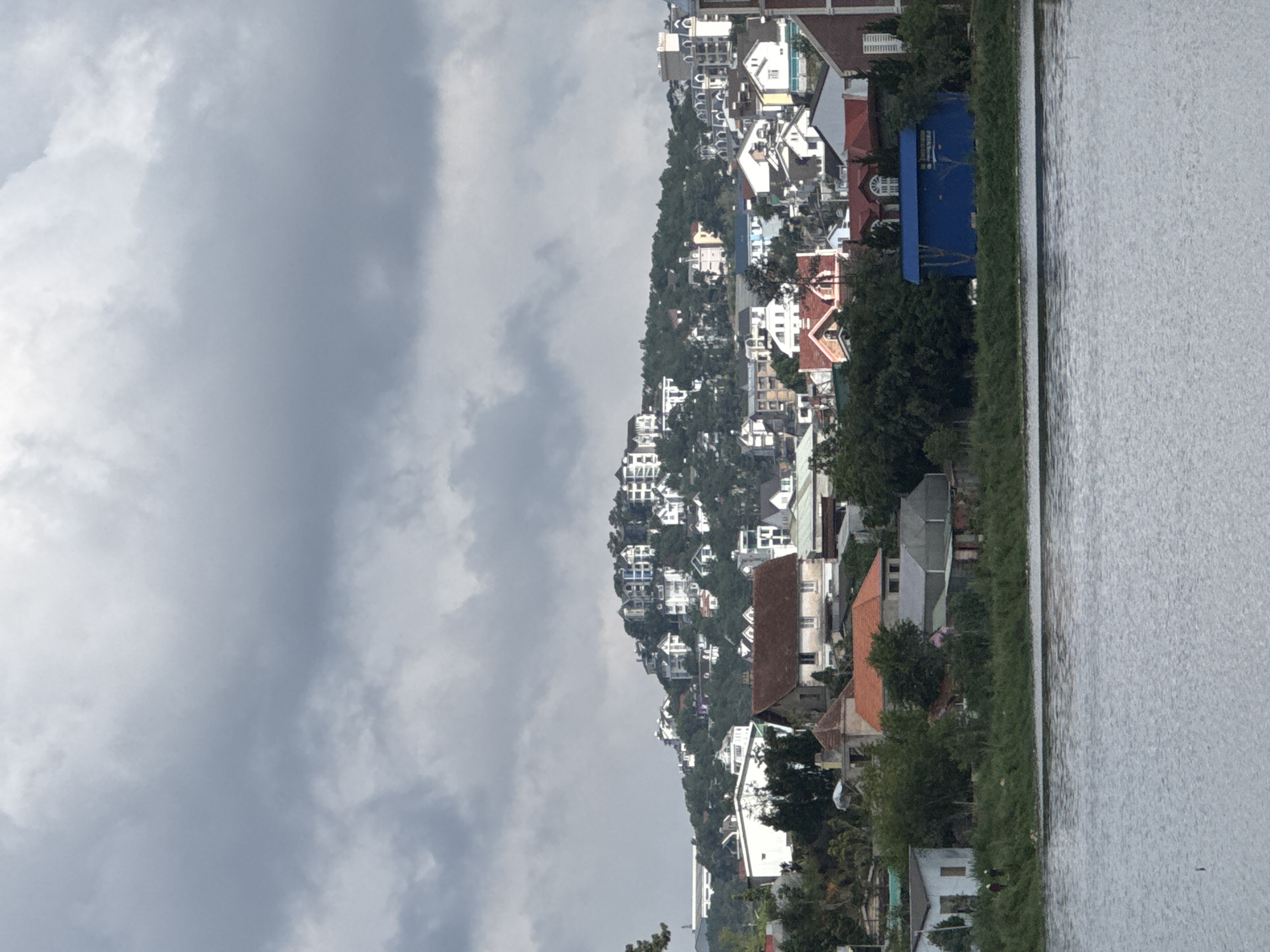 This media file captures a panoramic landscape scene, rotated vertically, depicting a hillside community beside a body of water under a cloudy sky.
The prominent feature of the image is a densely populated hillside, climbing from left to right (when viewed correctly). Numerous buildings, mostly houses, are constructed along the slopes, characterized by their varied architectural styles. Many houses are white or light-colored with distinct roofs, some red or brown, and others appear to be multi-story. Green trees and dense vegetation are interspersed among the buildings, covering parts of the hill and softening the urban sprawl.
In the foreground/right side of the image, a calm body of water extends across the frame. Its surface shows subtle ripples, reflecting the overcast sky. A narrow strip of green grass and foliage separates the waterfront buildings from the water itself. Among these waterfront structures, a striking blue building with a blue roof and distinct rounded window features prominently.
The sky is heavily overcast with various shades of grey clouds, suggesting a cloudy day, possibly before or after rain, and obscuring any direct sunlight. No people or specific activities are visible in the image, presenting a tranquil, static view of the landscape. Given the context of Lâm Viên - Đà Lạt, Vietnam, the architecture and hilly terrain are characteristic of this mountainous region. No visible text is present in the image.