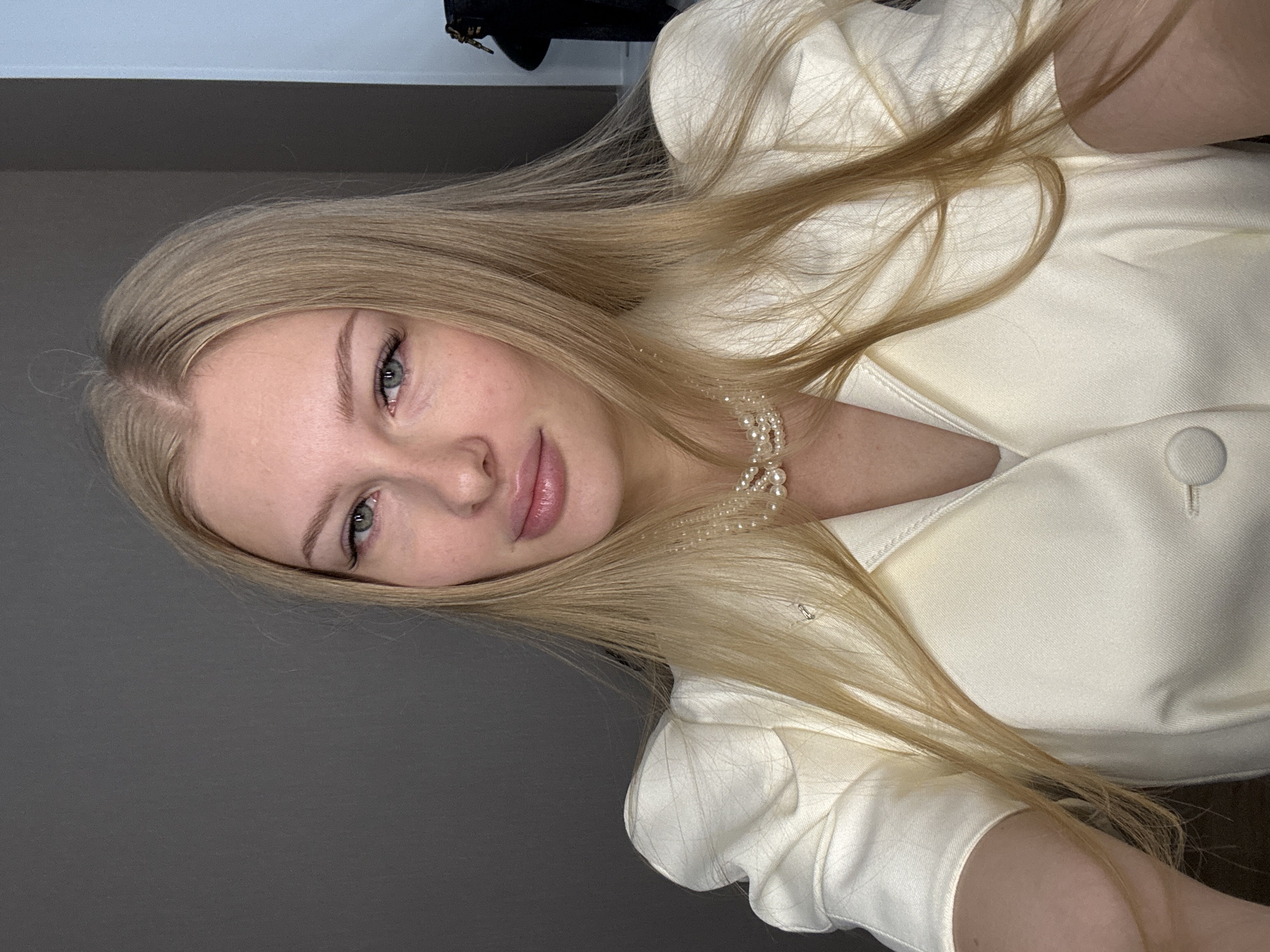 The image is a close-up, selfie-style shot of a young Caucasian woman with long blonde hair and light blue eyes. She is wearing a cream-colored blazer with puffed sleeves and a pearl necklace. Her makeup appears to be natural, with emphasis on her eyes and lips. The background is a plain grey wall, suggesting an indoor setting. There is a hint of a dark object in the upper right corner, possibly a bag or clothing item. The overall impression is one of casual elegance. The location is not specified in the image, and therefore it is not possible to determine if it is in Perm, Russia.