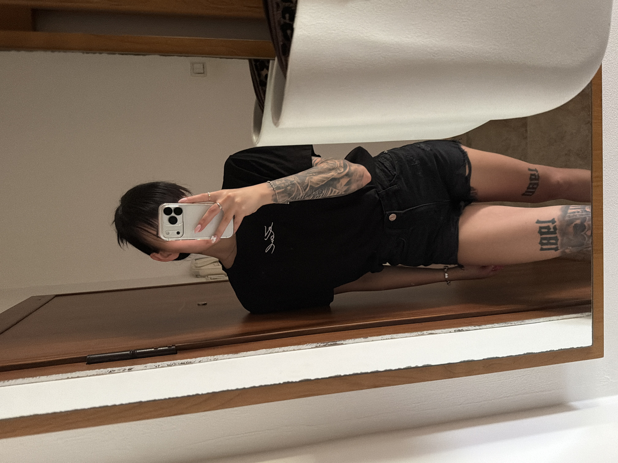 A young adult, likely female, stands in front of a mirror, taking a selfie with a light-colored smartphone held in her right hand, obscuring her face. She wears a black short-sleeved t-shirt with a white script-like logo on the chest, and black distressed denim shorts. Her left arm (visible on the right side of the image) is covered with a full sleeve tattoo in grey tones. Both thighs, visible above the knee, feature large, blocky numeral tattoos (e.g., "1981" on the person's right thigh and "1989" on the person's left thigh, both reversed in the mirror's reflection). She has short, dark hair with bangs, and wears multiple rings on her right hand, along with a silver-colored beaded bracelet on her left wrist. Her fingernails are painted light pink.
The mirror reflects a room with light-colored walls and a light brown wooden door with a dark metal hinge on the left. A small, white rectangular object, possibly a light switch or outlet, is visible on the wall in the background. A large, white cylindrical object (possibly a lamp base or decorative element) is positioned on the right side of the reflection, partially obscuring a patterned surface behind it. The mirror itself has a thin, light-colored wooden frame. The lighting is artificial and indoor, casting a soft, warm glow. The image does not contain any discernible visual cues or specific details that would allow for the identification of its location as Plovdiv, Bulgaria.