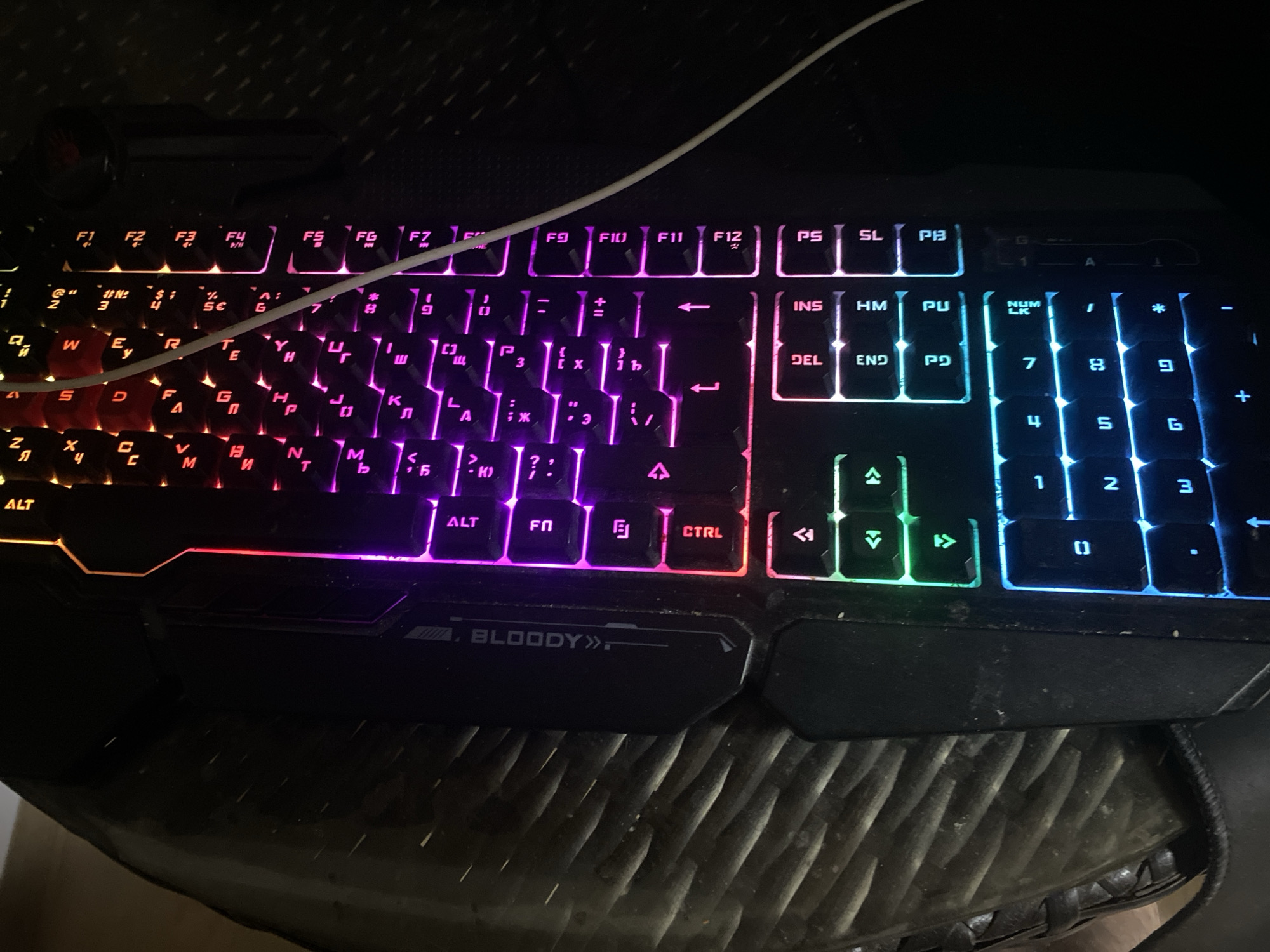 The image shows a close-up of a glowing mechanical keyboard, illuminated by a vibrant RGB lighting system. The keyboard is a Bloody brand, as indicated by the logo on the left side. The keys are lit in a variety of colors, including purple, pink, red, and blue, creating a striking visual effect. The keys themselves appear to be standard QWERTY layout, with some function keys labeled at the top and navigation keys along the bottom. The keyboard is resting on a dark, textured surface, possibly a desk or a woven mat. The overall impression is one of a modern, gaming-oriented keyboard. The location of Bol'shoe Zaborod'e, Russia is not discernible from the image itself, as there are no visual cues related to this specific geographic area.
