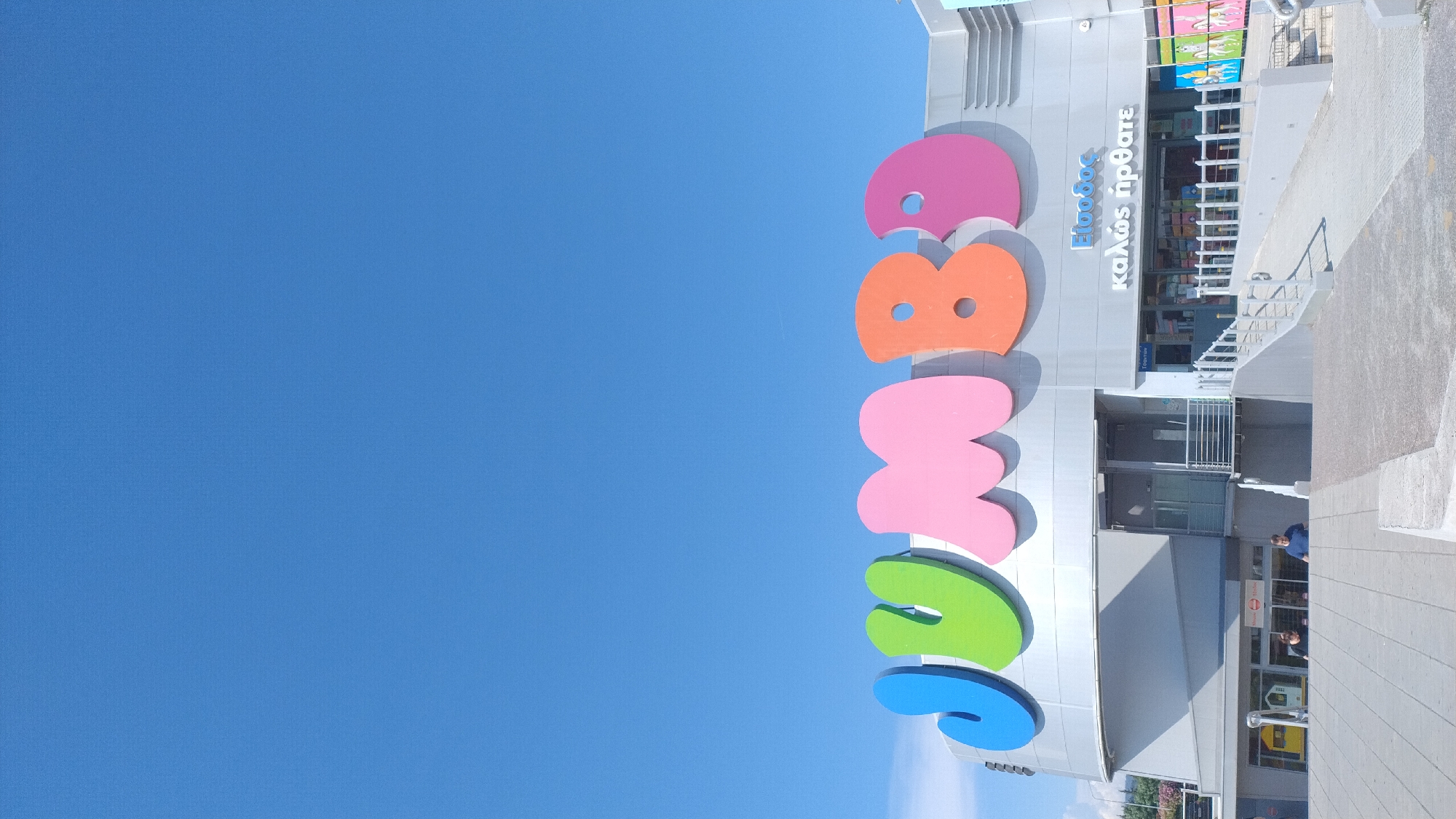 A vertical, eye-level, exterior shot depicts the facade of a Jumbo store under a clear blue sky in Pylaia Municipal Unit, Greece. The prominent, colorful "JUMBO" logo, with letters in blue, green, pink, orange, and purple, spans the upper portion of the building. Below the logo, a gray, modern building with multiple entrances and a ramp leading up to one of the entrances is visible. Two people are standing near the lower entrances, one near a yellow doorway on the left, and another in a blue shirt in the center. The ground in front of the building is paved with gray concrete, with a concrete barrier in the foreground. The overall impression is a bright, sunny day at a large retail establishment.