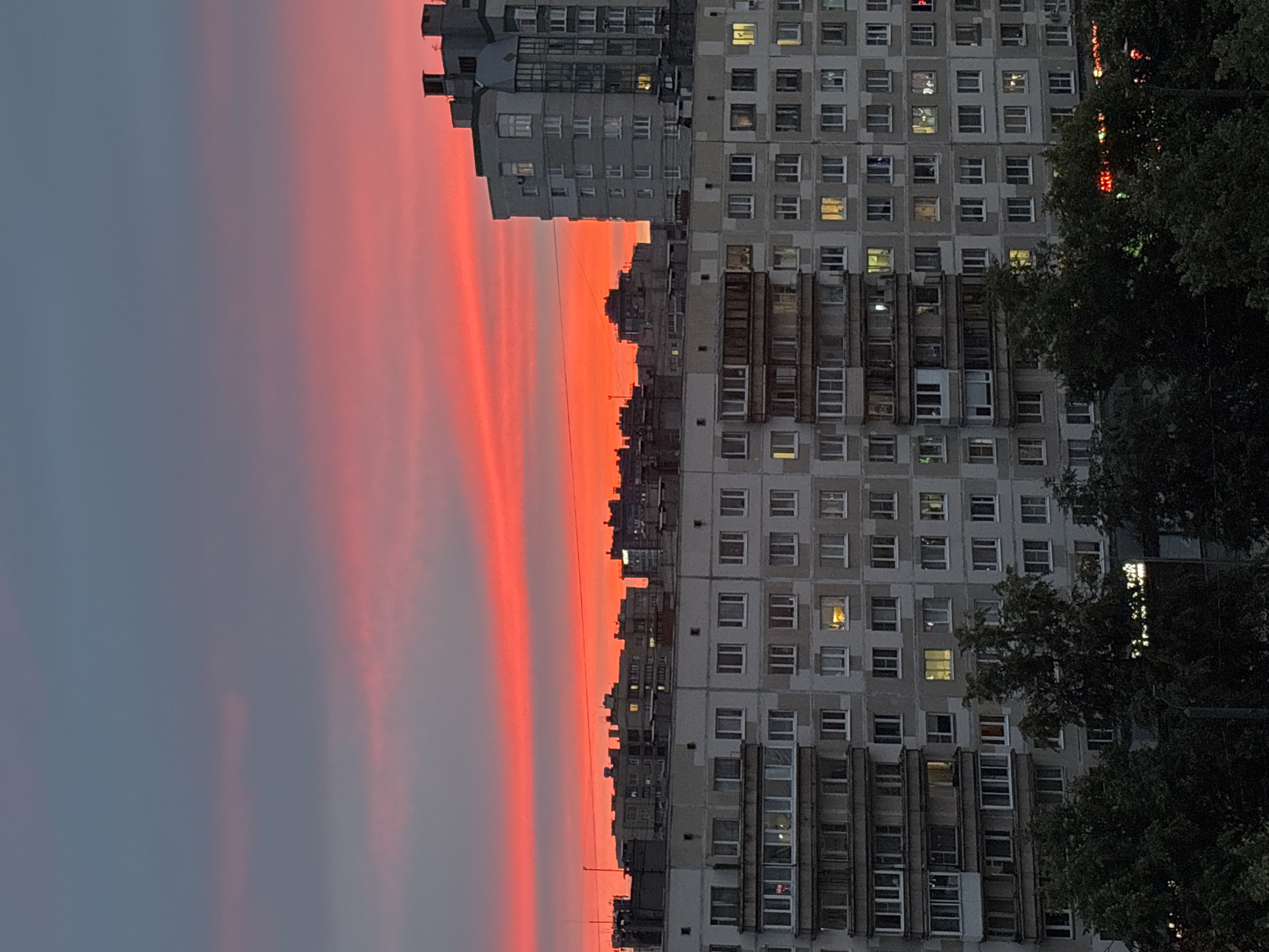 The image captures a dramatic sunset over a city skyline. The sky is painted with vibrant streaks of red and pink, transitioning into a muted blue-grey at the top. In the foreground, multi-story apartment buildings dominate the scene. These structures are characterized by their uniform windows and repetitive architectural style, typical of Soviet-era housing blocks. Some windows are illuminated, suggesting residents are home. Trees with dark foliage are visible at the bottom of the frame, partially obscuring the lower levels of the buildings and adding a natural element to the urban landscape. The overall impression is one of a twilight urban environment under a spectacularly colored sky. The location is identified as Unknown, Russia.