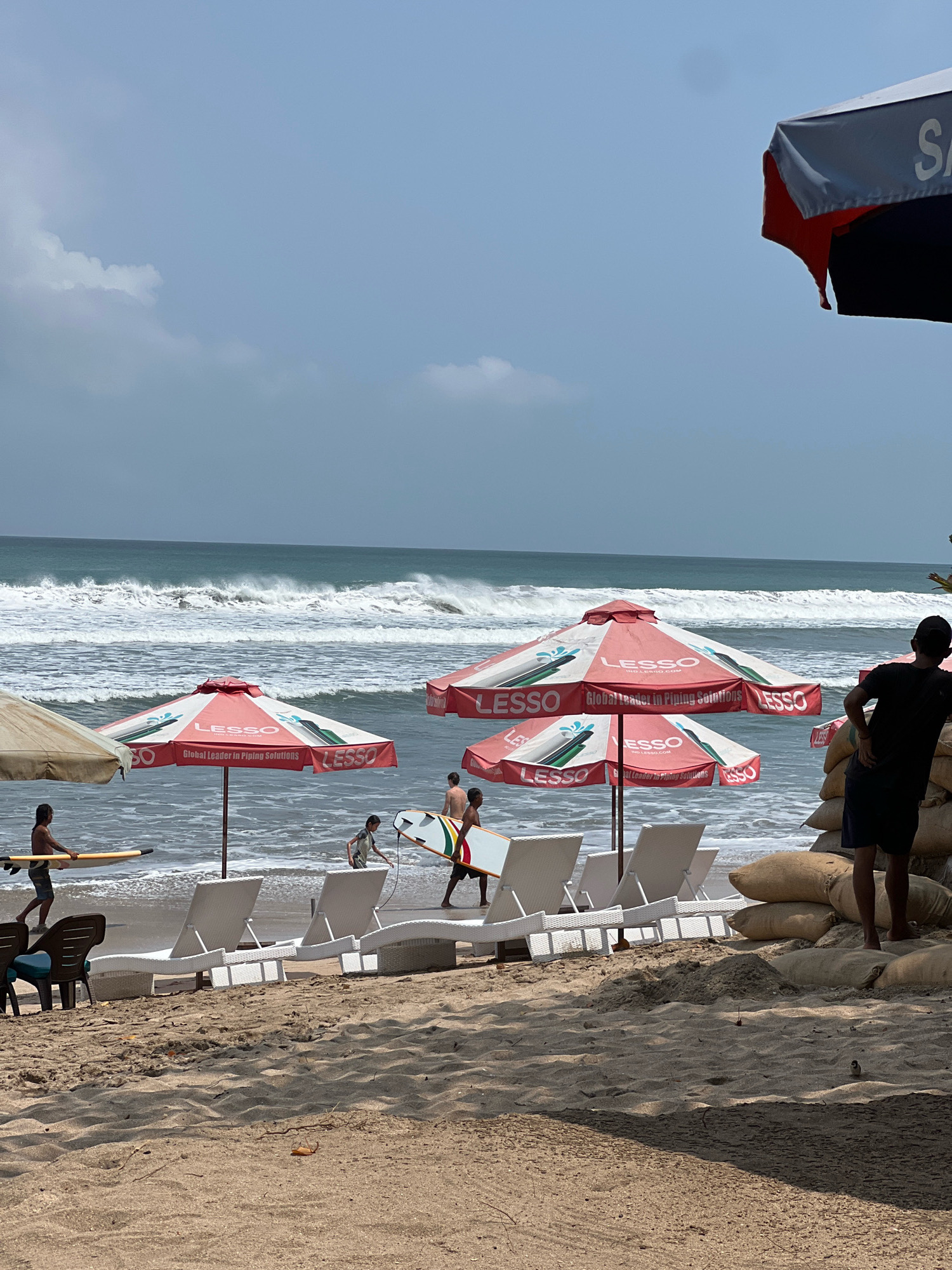 In the city of Kuta, Indonesia, this image captures a beach scene with the ocean in the background. The water is a deep blue with white waves crashing. Several beach umbrellas are visible, some red and white with the "LESSO" logo and a slogan about piping solutions, and one partially visible umbrella on the far right with red and black fabric. Beach chairs, mostly white and wicker, are arranged on the sand. In the foreground, there are sandbags piled up. Several people are present: one individual in the foreground is wearing a black t-shirt and dark shorts, facing away from the camera and looking towards the ocean. A few other individuals, including children, are closer to the water, some carrying surfboards. One person is walking in the water with a surfboard. The overall environment is sunny with a slightly cloudy sky.