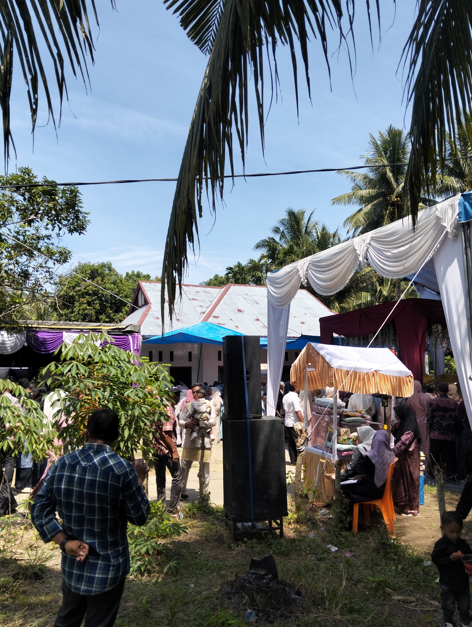 This is a medium shot, outdoor photograph of a gathering in Pulonga, Indonesia. The scene appears to be an event, possibly a wedding or celebration, with several people dressed in formal attire. The focal point of the image is a man in a blue and green plaid shirt standing with his back to the camera, facing a crowd and several tents. One tent is white with gold trim, and another is draped with white and purple fabric. A large black speaker is visible to the left of the man. In the background, a building with a corrugated metal roof and a blue tarp is visible, surrounded by lush green trees and palm fronds. The sky is clear and blue, indicating a sunny day. The ground is covered in grass and dirt, with some scattered debris. A small child in a black shirt is visible on the right side of the frame.