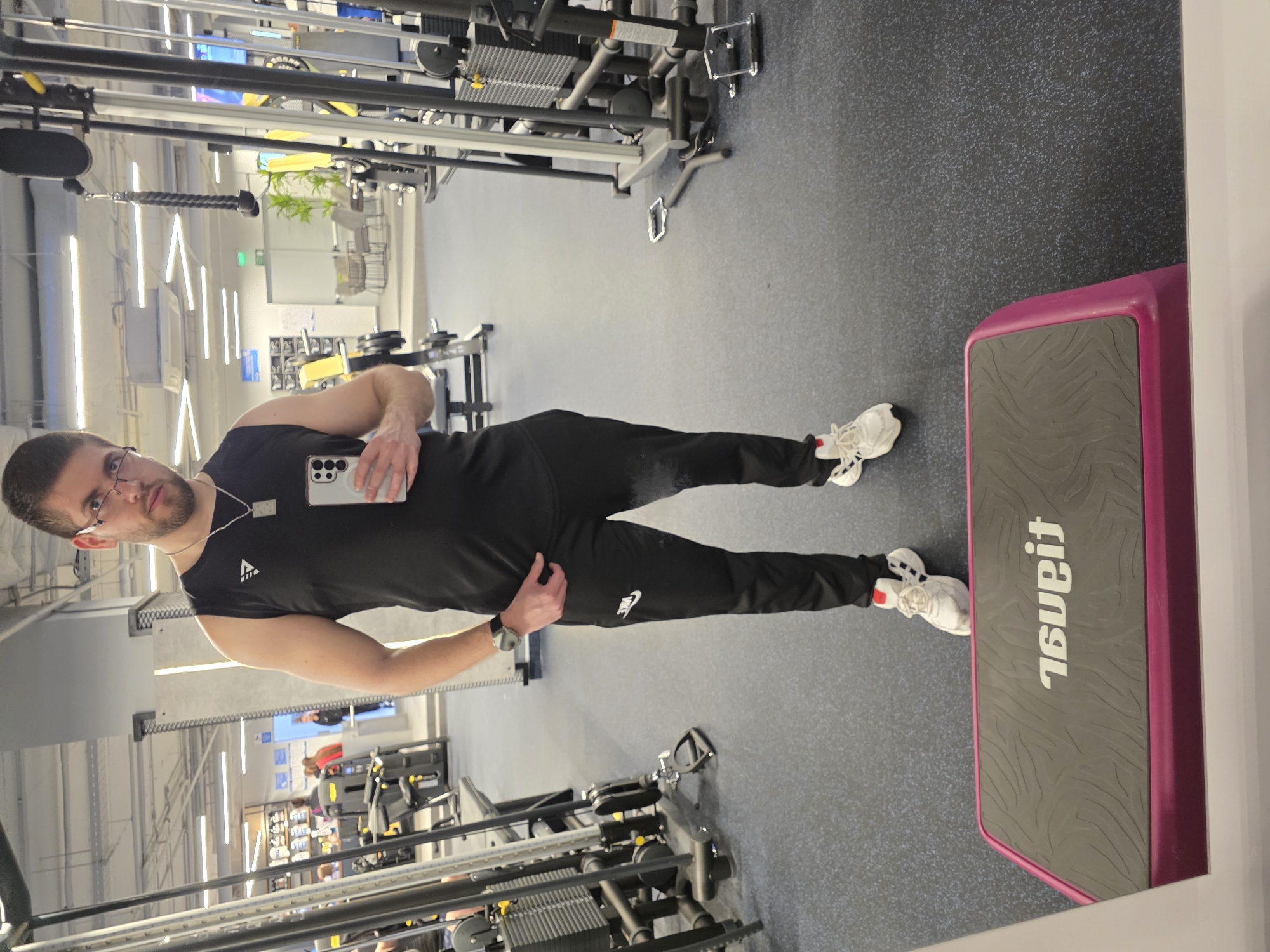 A man is standing in a gym. He is wearing a black tank top, black sweatpants, and white sneakers. He is holding a cell phone in his left hand. He also wears glasses, a watch, and a necklace. Behind him, there are various pieces of gym equipment, including weight machines. To his right is a step platform with the words "Planet" printed on it in reverse. The gym has a grey floor and bright overhead lighting. The location is most likely in a fitness center somewhere in an urban area. No verifiable information can pinpoint the gym to Plovdiv, Bulgaria.