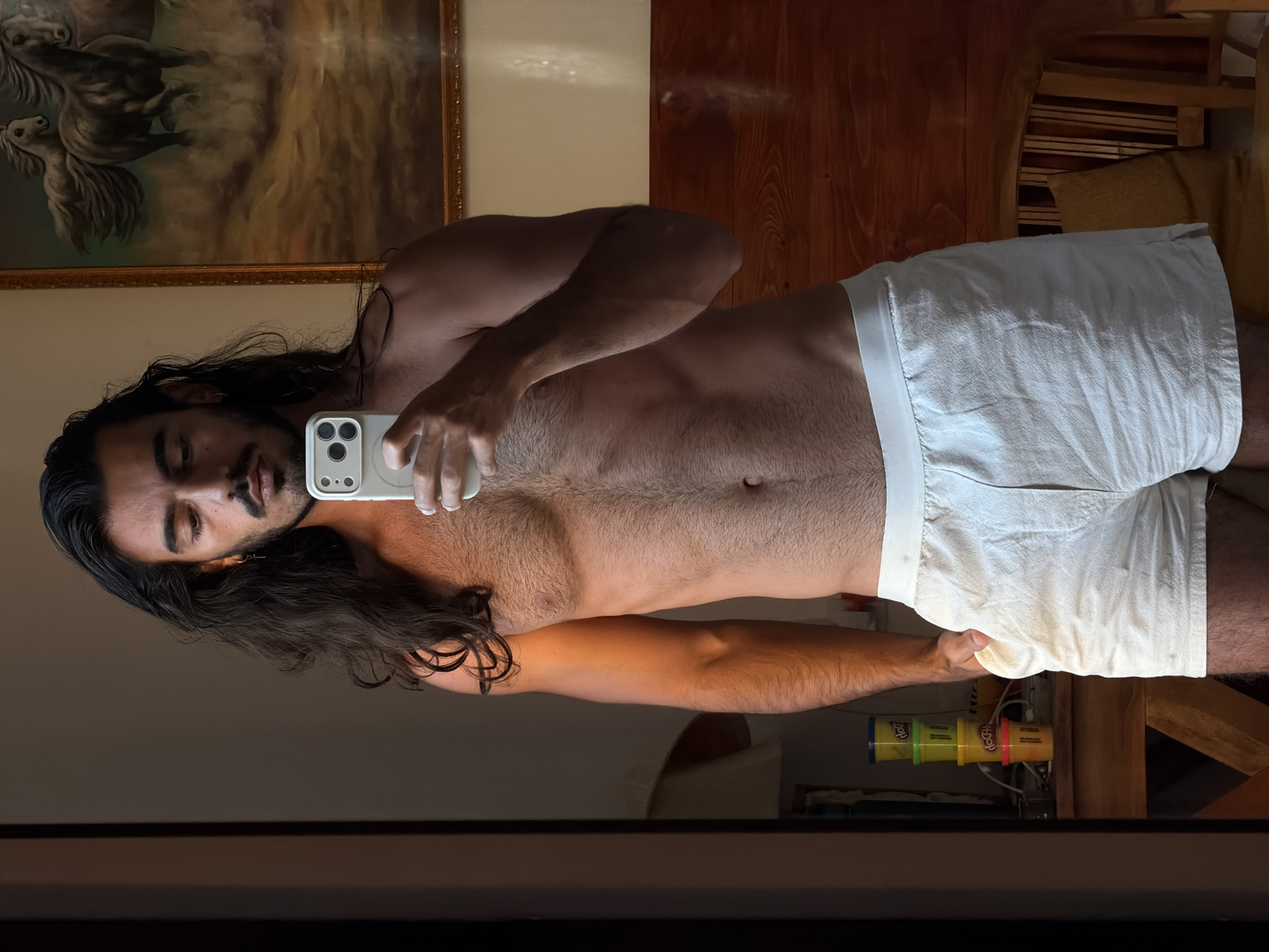 A shirtless man with long dark hair and a mustache takes a selfie in a mirror while wearing white shorts. He is holding a phone in his right hand. Behind him, there is a painting of horses on the wall and wooden floors. Underneath his shorts and arm, there are some containers of Play-Doh. It is not possible to ascertain the location in the city of North Kuta, Indonesia.