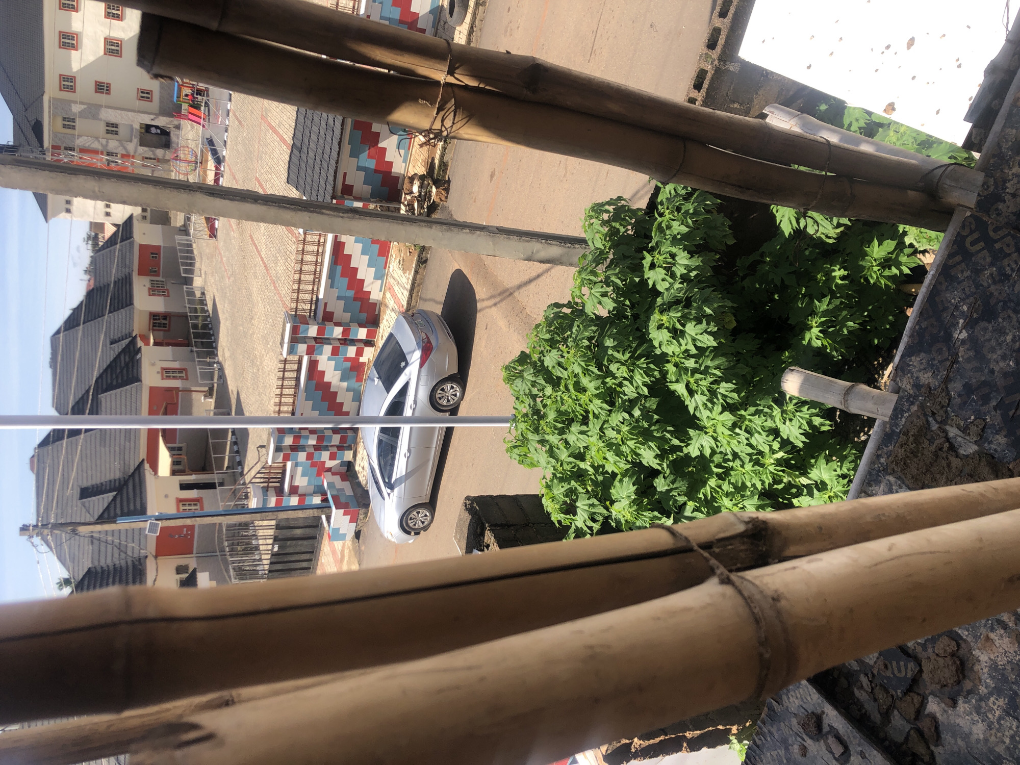 This is a photograph taken from a slightly elevated vantage point, looking down and across a residential area. The foreground is dominated by the texture of bundled bamboo poles, suggesting a construction or scaffolding element. Beyond the bamboo, a silver sedan is parked on a paved road. A vibrant green bush with large leaves occupies the middle ground. In the background, several modern residential buildings with dark pitched roofs and light-colored walls with red and white accents are visible. A colorful playground structure with slides and climbing equipment is present in the courtyard of one of the buildings. The scene appears to be set in a bright, daytime environment with clear skies. The context provided indicates the location is in the city of Gura Top, Nigeria.