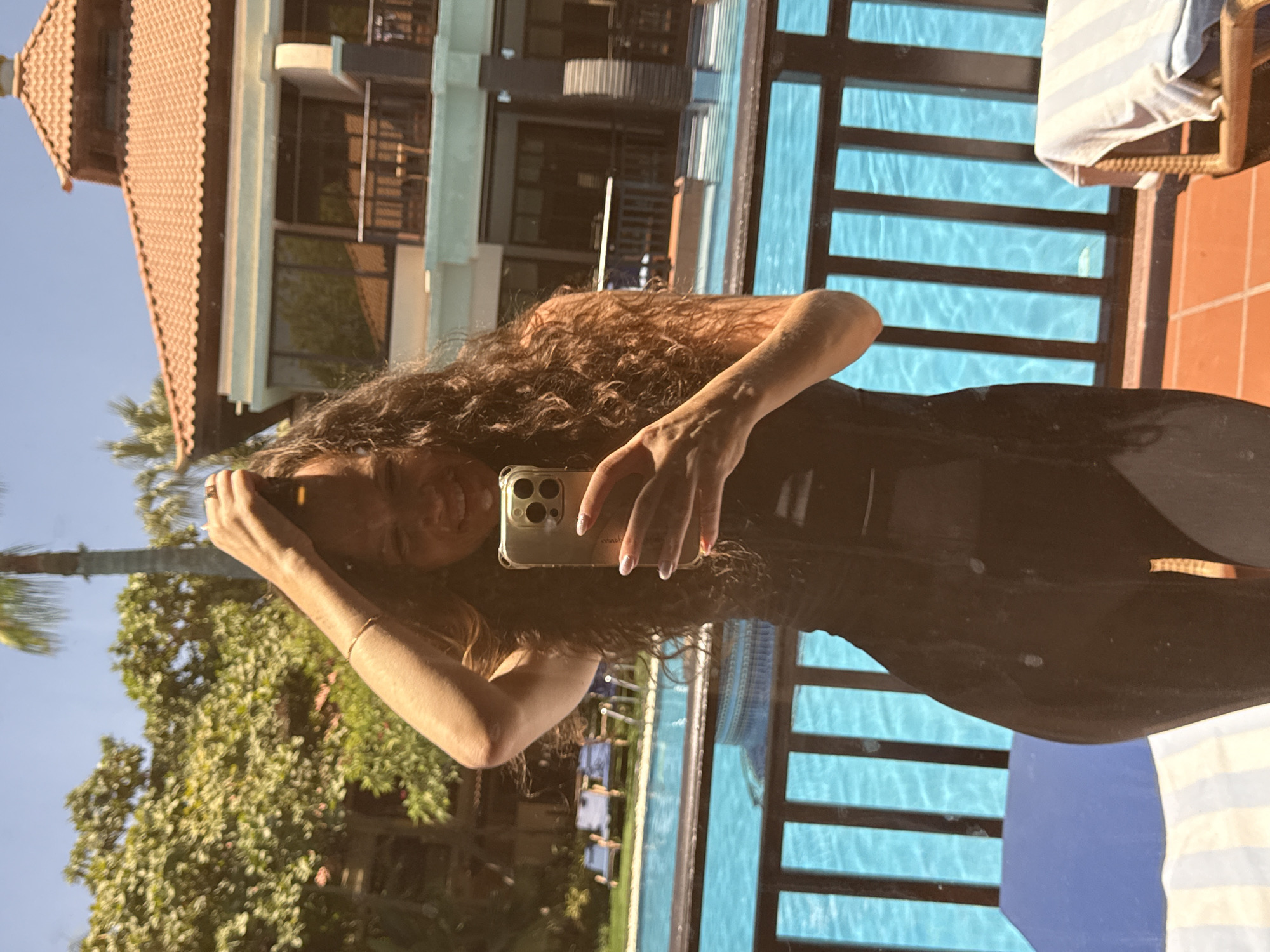 A woman with long, curly brown hair is taking a mirror selfie in what appears to be a resort or hotel. She is wearing a dark, form-fitting outfit, possibly a swimsuit or athletic wear. Her right hand is holding a gold smartphone, and her left arm is raised, with her hand resting on her head. She is smiling at the camera.
Behind her, a bright blue swimming pool is visible, bordered by a dark wooden railing. In the background, there is a building with a terracotta-tiled roof and balconies, suggesting a tropical or Mediterranean architectural style. Lush green trees and a palm tree are also present, indicating an outdoor, possibly sunny, environment. The ground is tiled with orange, square tiles. There are lounge chairs with blue cushions and striped towels by the pool. The overall scene suggests a vacation or leisure setting. There is no specific information to pinpoint the location to Dubai, United Arab Emirates.