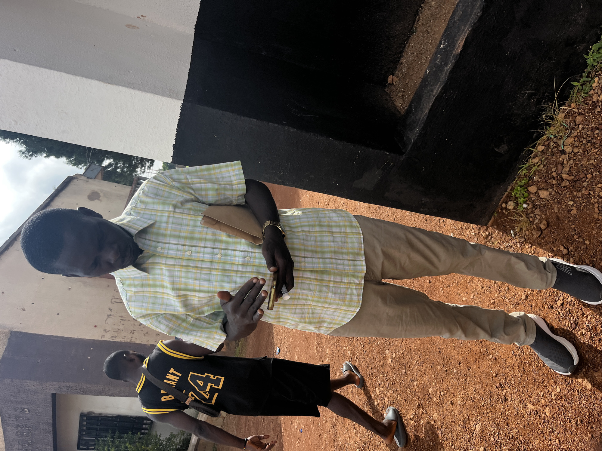 In Gura Top, Nigeria, on a dirt path beside a building with a light-colored wall and a black base, two men are visible. The man in the foreground, wearing a light green and white checkered short-sleeved shirt, tan pants, and gray slip-on athletic shoes, holds a light brown wallet in his left hand and a cell phone in his right hand, which he is looking at. A man in the background walks away, wearing a black sleeveless jersey with yellow lettering and numbers "BRYANT 24", black shorts, and gray slide sandals. The ground is covered in reddish-brown dirt and small rocks, with some sparse green vegetation. The sky is overcast.