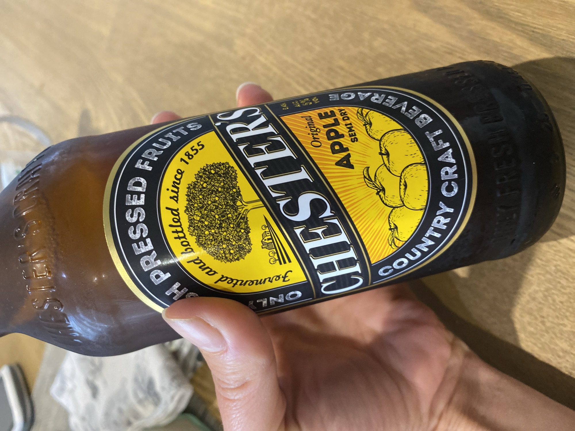 A hand holds a dark brown glass bottle of cider. The bottle has a round label with a yellow background and black text. The label states "CHESTERS" prominently in the center, with "ORIGINAL APPLE SEMI DRY" below it. A graphic of an apple tree and several apples is also visible on the label. The bottle is cool and appears to have condensation on it. The background is a light-colored wooden surface. The image is taken from a close-up perspective, focusing on the bottle and the hand.