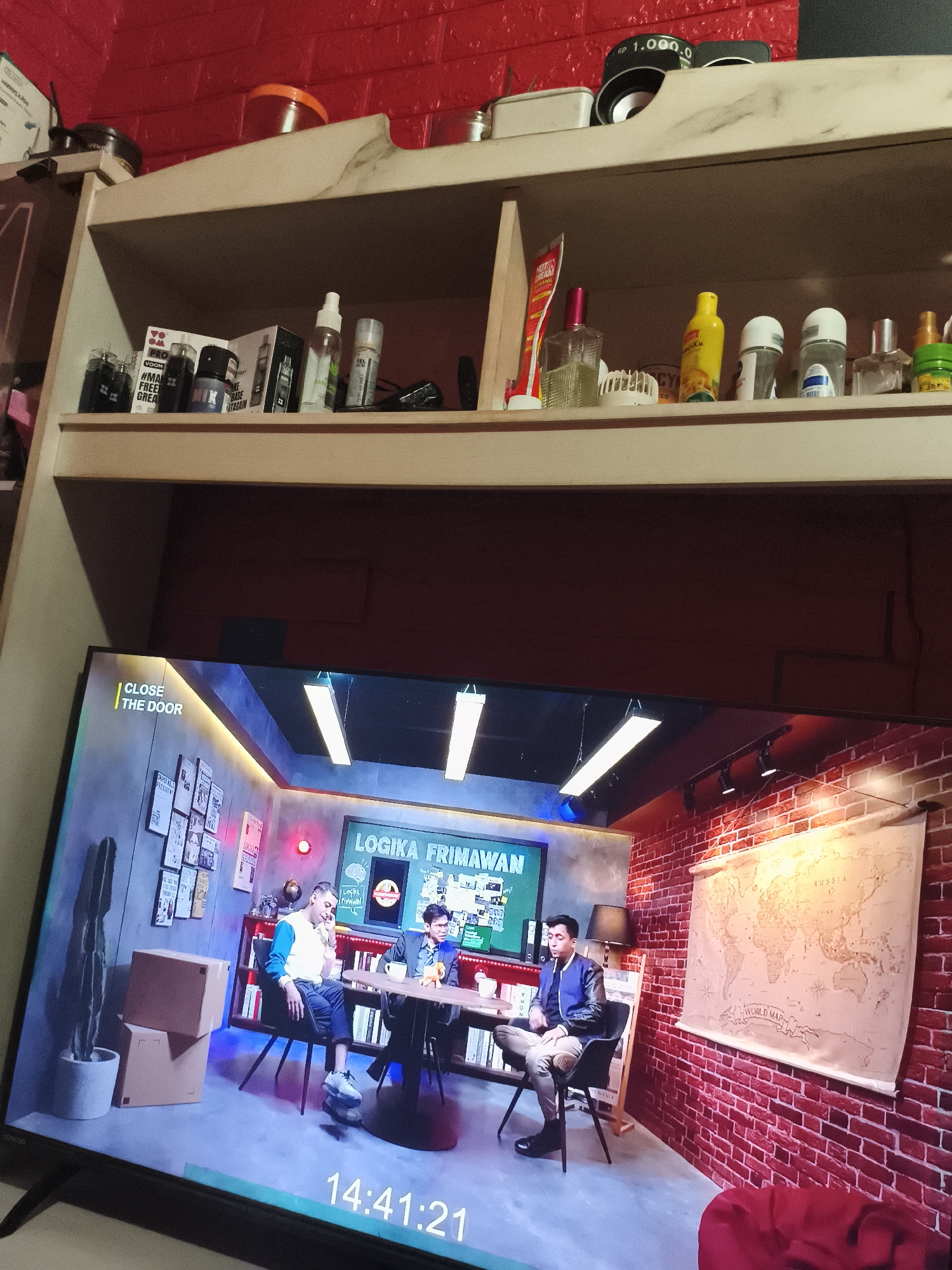 The image depicts a shelf with various items on top, positioned above a television screen displaying a talk show titled "Logika Frimawan" with three people seated at a table. The show features a set decorated to resemble a brick-walled room with a world map. The clock on the TV displays the time as 14:41:21. The shelf contains a variety of personal items including cosmetics, skincare products, a speaker, and various containers. The location can be inferred to be in Depok, Indonesia based on the language and the context of the television program.
