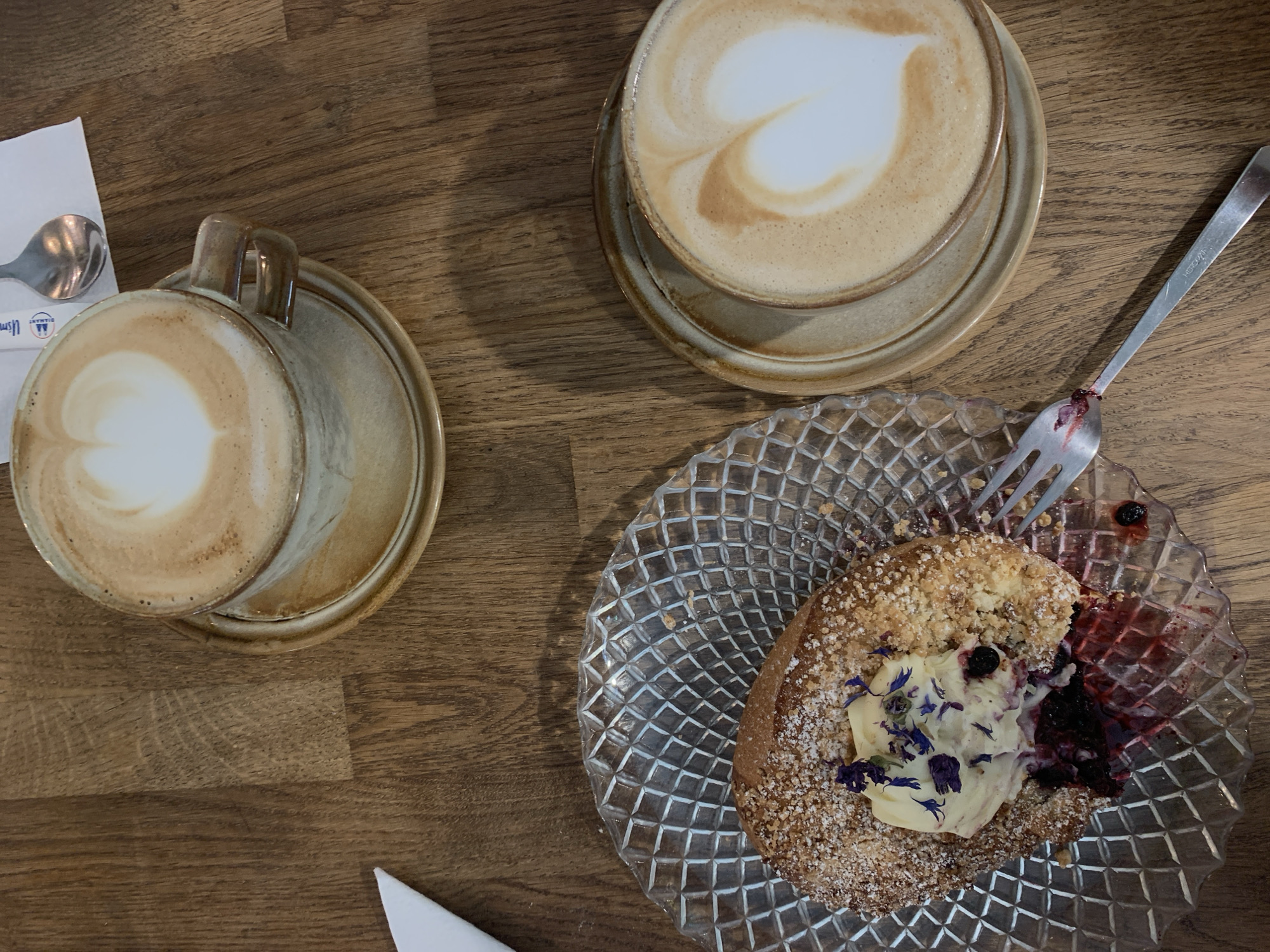 The overhead shot captures a wooden table setting with two lattes and a pastry. The lattes, served in ceramic cups with latte art in the shape of hearts, sit on saucers. A fork, with remnants of a dark berry sauce, rests beside a textured glass plate. On the plate is a pastry topped with cream, blueberries, and small purple flowers, with a berry filling visible. The scene suggests a cafe environment, possibly in Poznan, Poland, given the context. The lighting is warm, casting shadows on the wooden surface.